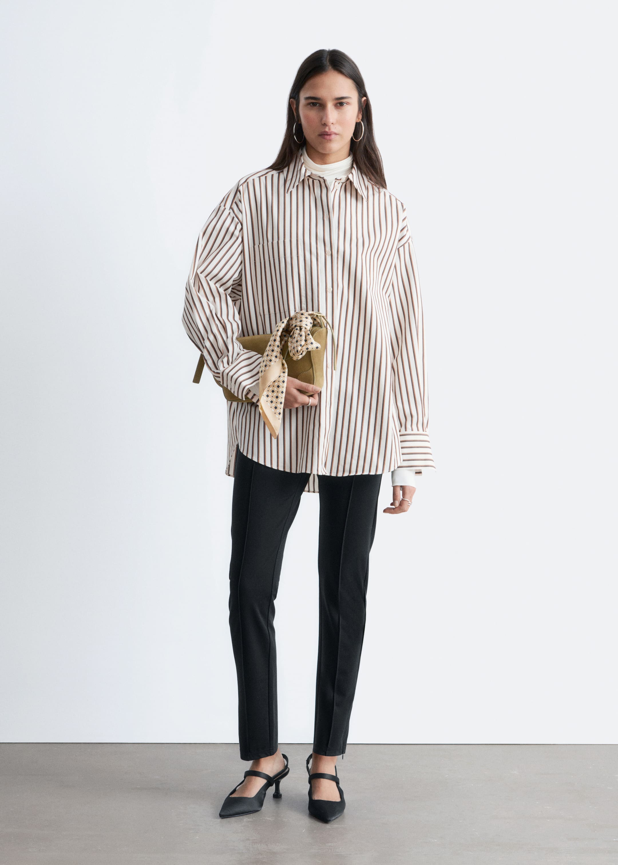 Zobacz większy obraz: Modelka nosi oversize'ową koszulę w paski, z kołnierzykiem typu turn-down, kieszonką na piersi i mankietami zapinanymi na guziki. Koszula jest założona na biały golf, a modelka trzyma oliwkową kopertówkę ozdobioną wzorzystą apaszką.