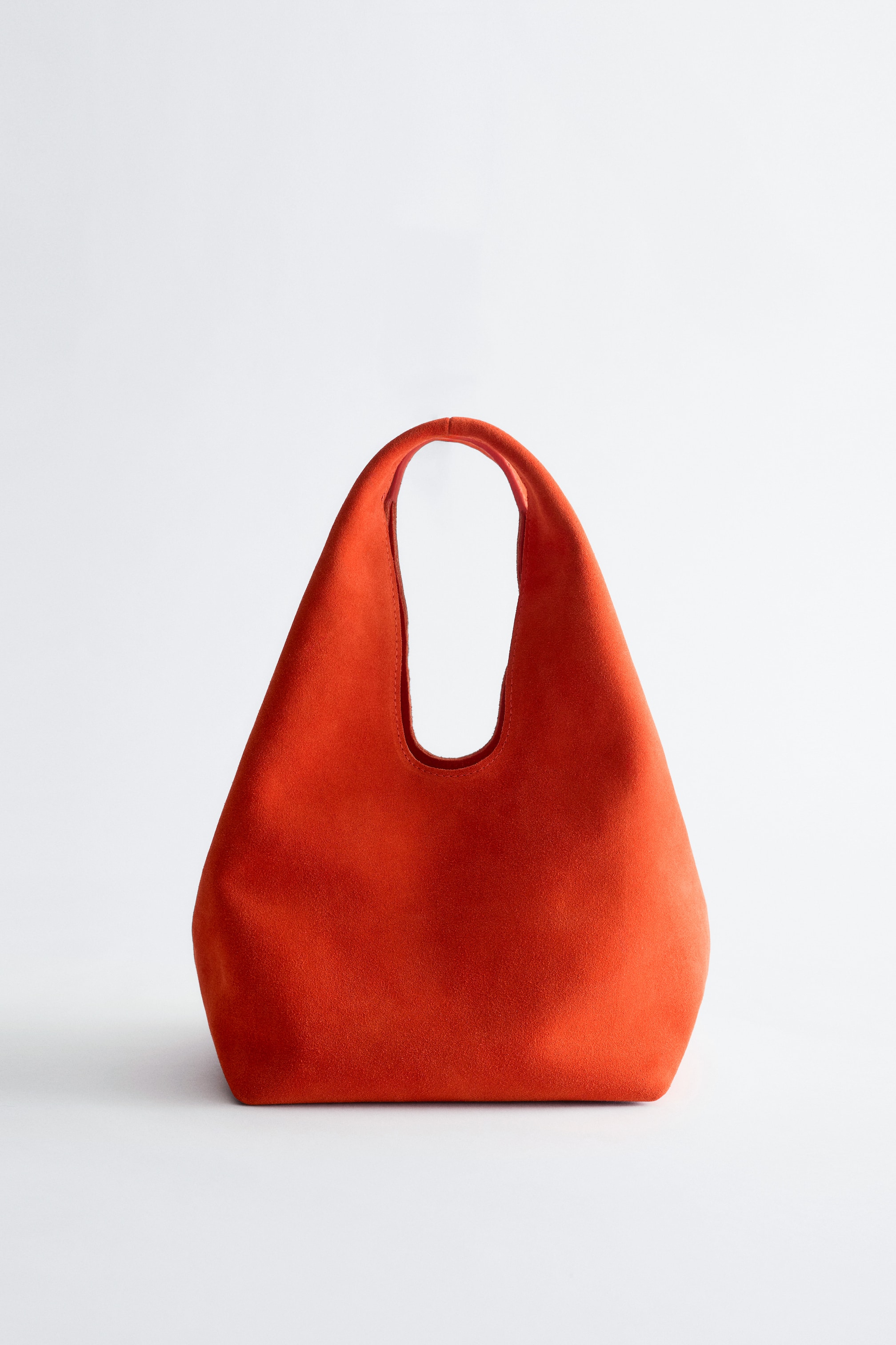 Ver imagem maior: Uma pequena mala tote laranja vibrante está virada para a frente, apresentando uma silhueta curva com uma textura suave semelhante a camurça. A sua alça integrada exibe detalhes dobrados na parte superior.