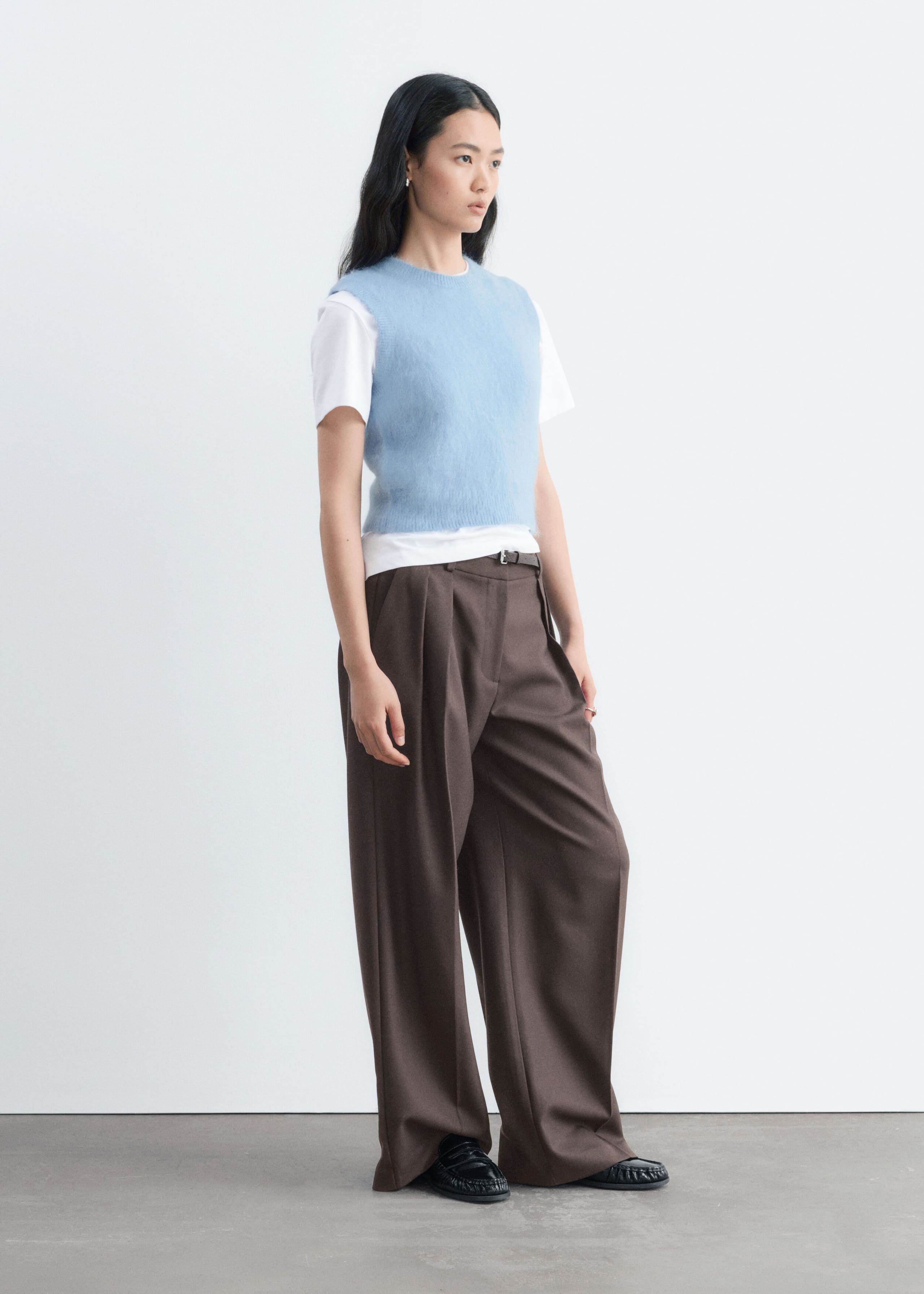 La modelo lleva un chaleco de mezcla de mohair de punto en azul claro, con textura afelpada, cuello redondo y bajo acanalado. El chaleco se lleva sobre una camiseta blanca de manga corta y se combina con pantalones marrones de pernera ancha.