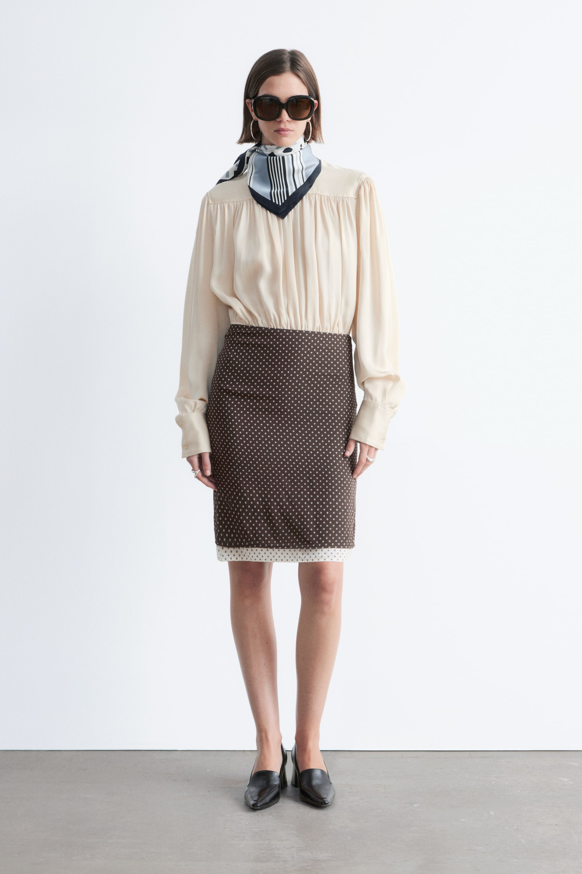 Visualizza immagine più grande: Una blusa color crema a maniche lunghe presenta uno scollo arricciato, maniche voluminose e polsini con bottoni. È infilata in una gonna a tubino marrone al ginocchio, caratterizzata da un motivo di piccoli punti chiari e sovrapposta a un capo bianco con un motivo a punti abbinato, visibile sull'orlo.