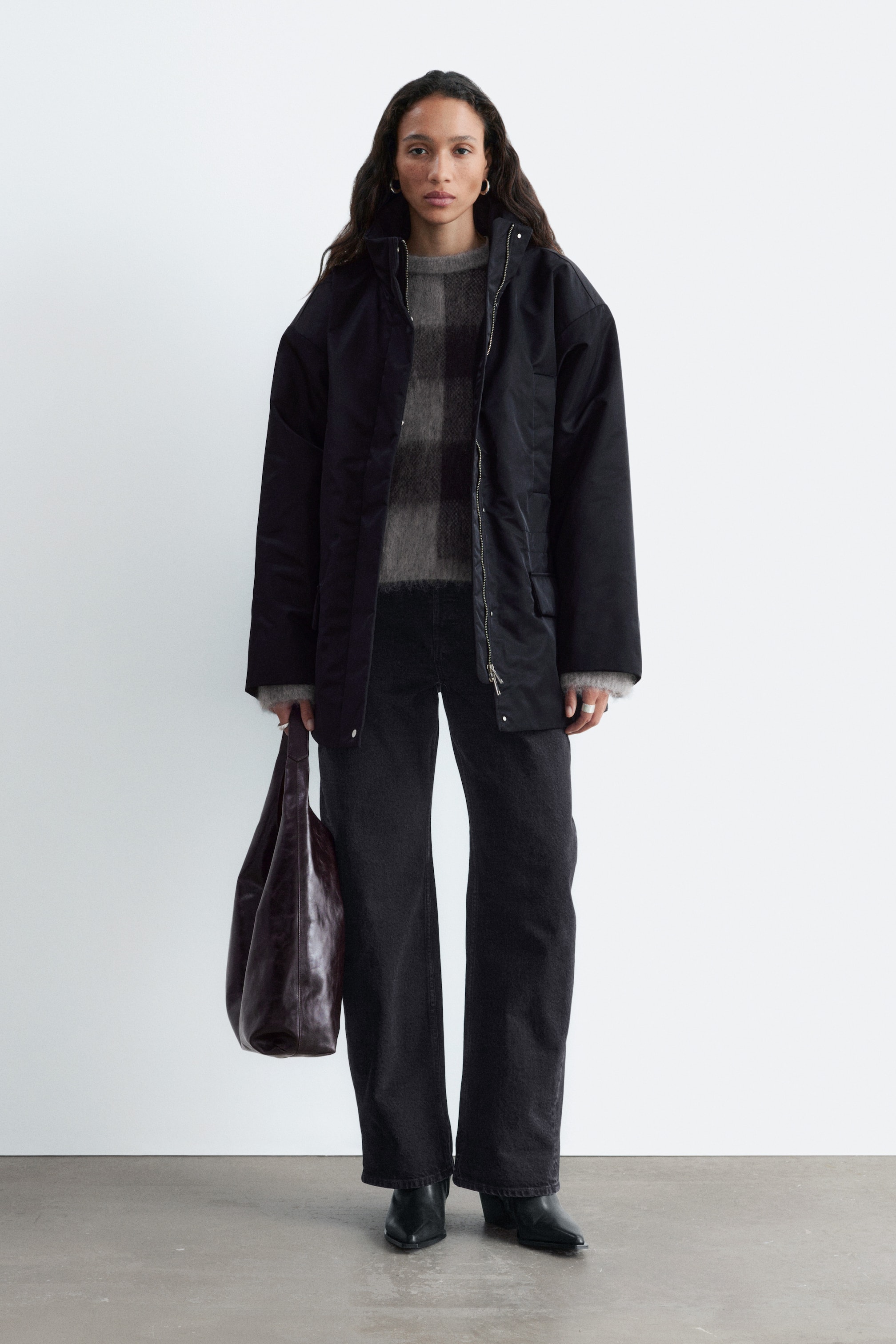 Größeres Bild anzeigen: Eine schwarze, gepolsterte Nylonjacke mit hohem Kragen und Reißverschluss wird vom Model offen getragen. Die Jacke ist über einem grau gemusterten Strickpullover und einer dunkelblauen Wide-Leg-Jeans getragen, dazu wird in einer Hand eine dunkle Lederhandtasche gehalten.