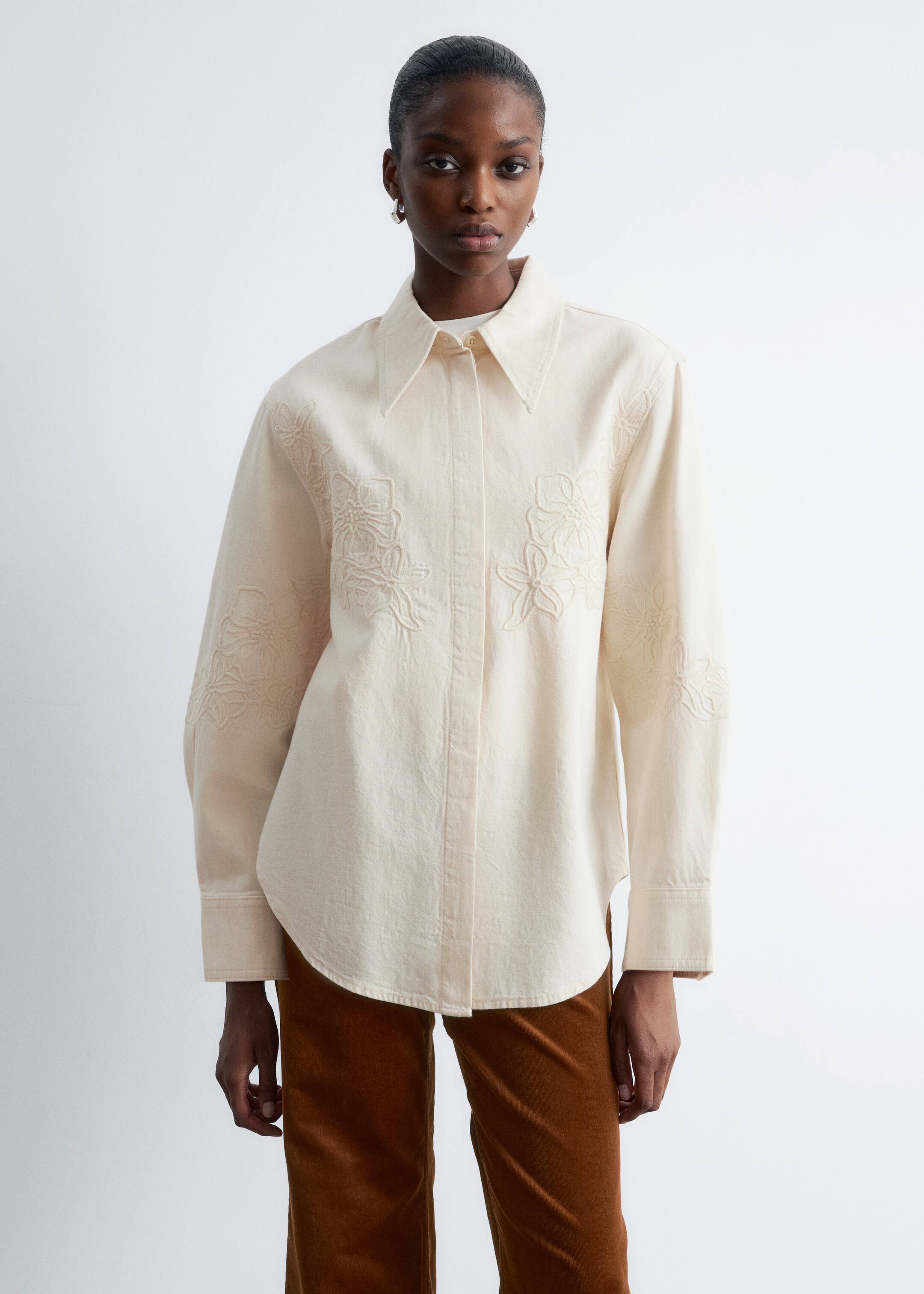Zobrazit větší obrázek: Modelka má na sobě krémovou džínovou košili s vyšívanými květinovými vzory na hrudi a horních rukávech. Volně padnoucí košile má klasický límec, dlouhé rukávy a zaoblený lem a je stylizována přes bílé spodní tričko s hnědými kalhotami.