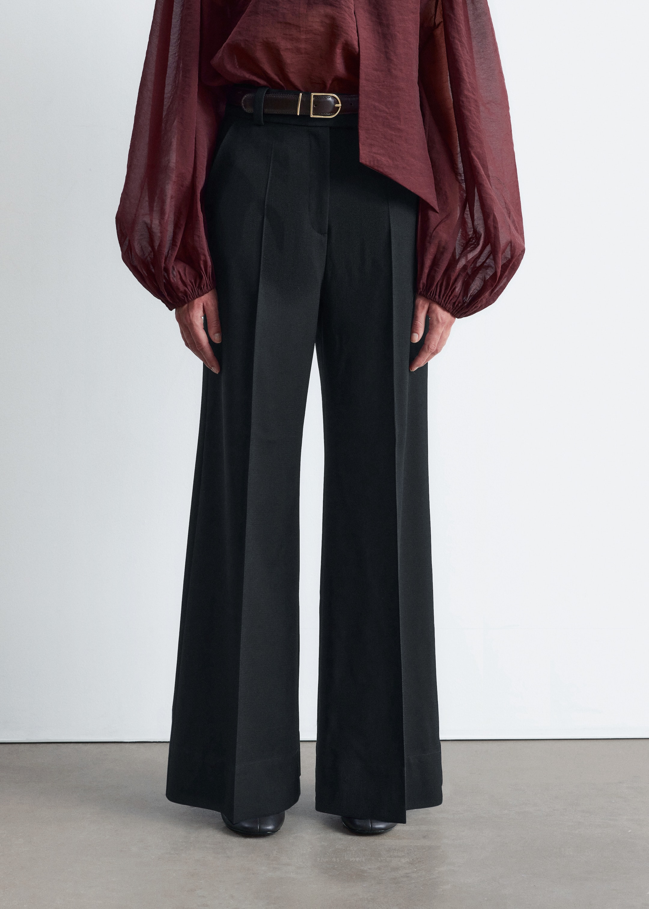 Visualizza immagine più grande: Pantaloni neri a gamba larga presentano una vestibilità a vita alta con una piega frontale prominente e un drappeggio liscio e fluente. Una cintura scura con fibbia color oro stringe la vita.