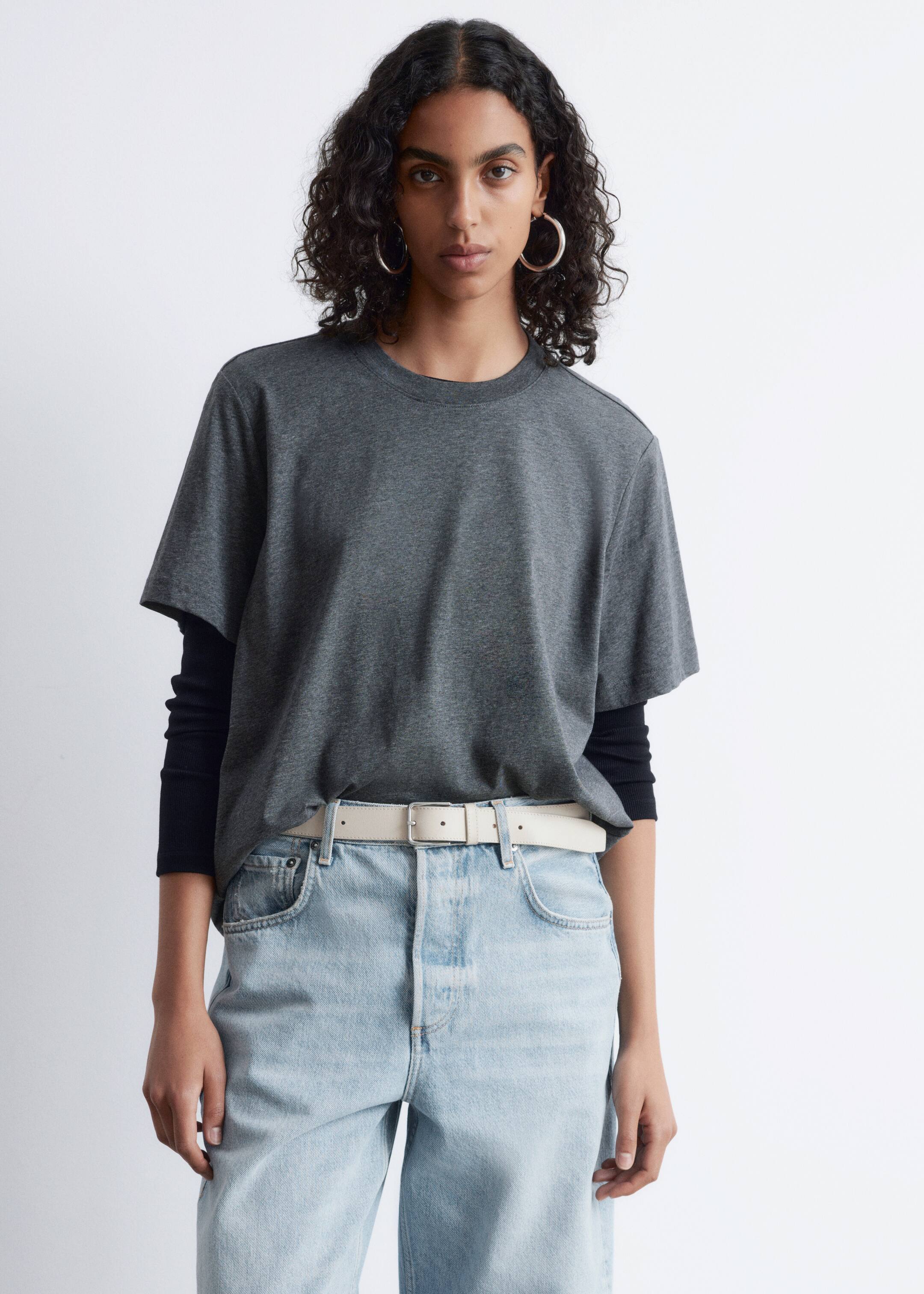 Vezi imaginea mai mare: Un tricou cu mâneci scurte și de o culoare gri închis cu croială lejeră este purtat de model. Este stratificat peste un top cu mâneci lungi negru și este parțial băgat în blugi decolorati, care sunt fixați cu o curea deschisă la culoare.
