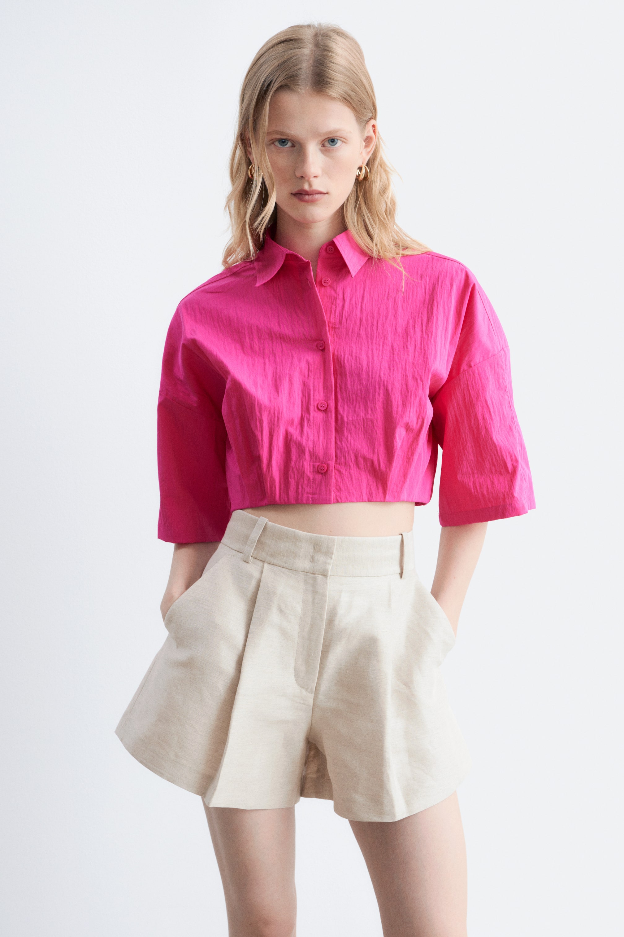 Ver imagen más grande: Una camisa corta texturizada de color rosa brillante, que presenta un cuello en punta, cierre frontal de botones y mangas anchas de tres cuartos de longitud, se muestra desde el frente. Unos pantalones cortos beige de talle alto con pliegues y bolsillos laterales completan el conjunto.
