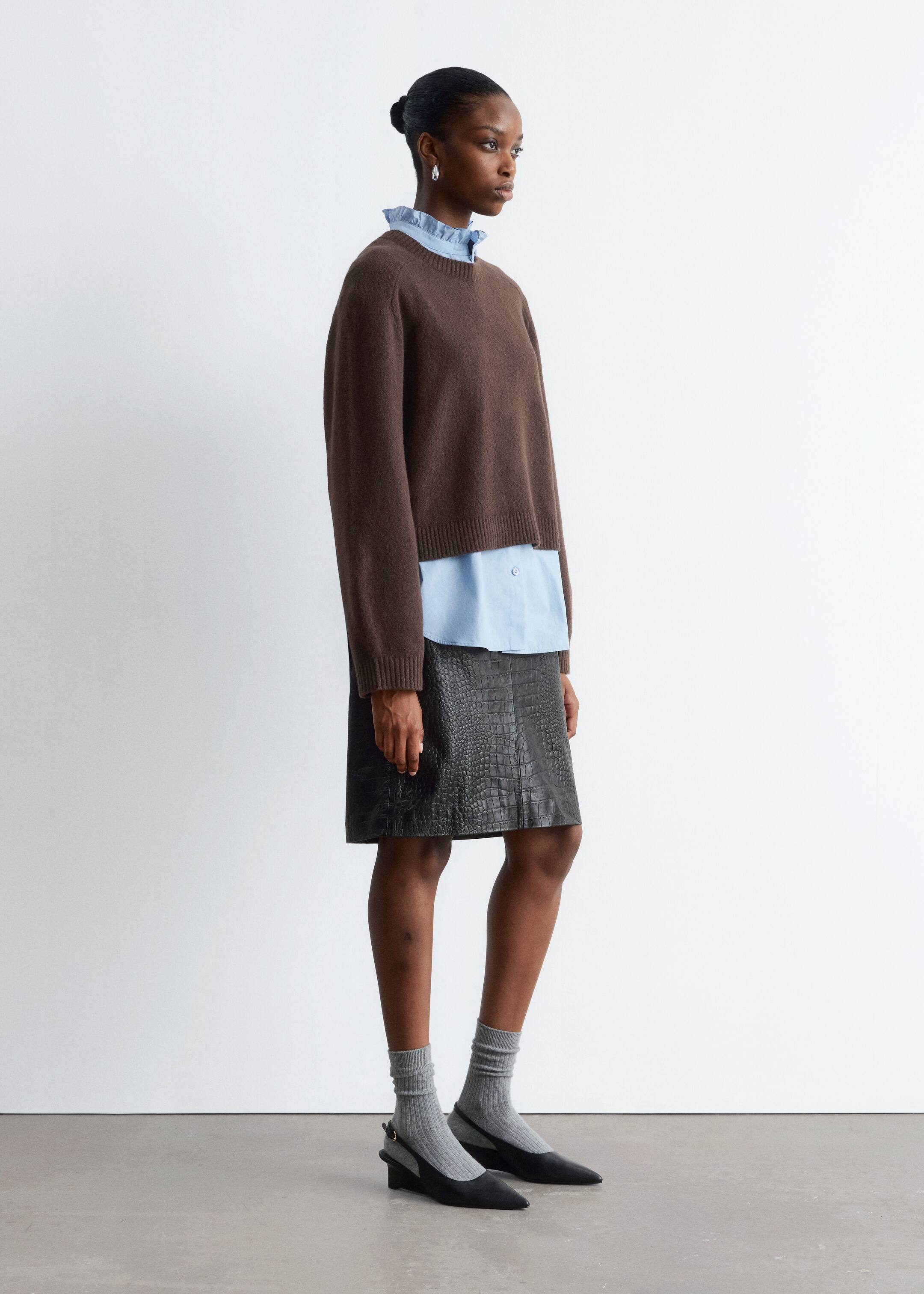 Grösseres Bild ansehen: Ein brauner Strickpullover mit Rundhalsausschnitt wird aus einem Seitenprofil gezeigt und hat eine entspannte, leicht verkürzte Passform. Er wird über einem hellblauen Hemd mit Kragen getragen, wobei der gerüschte Kragen und der Saum des Hemdes sichtbar unter dem Pullover hervorragen.
