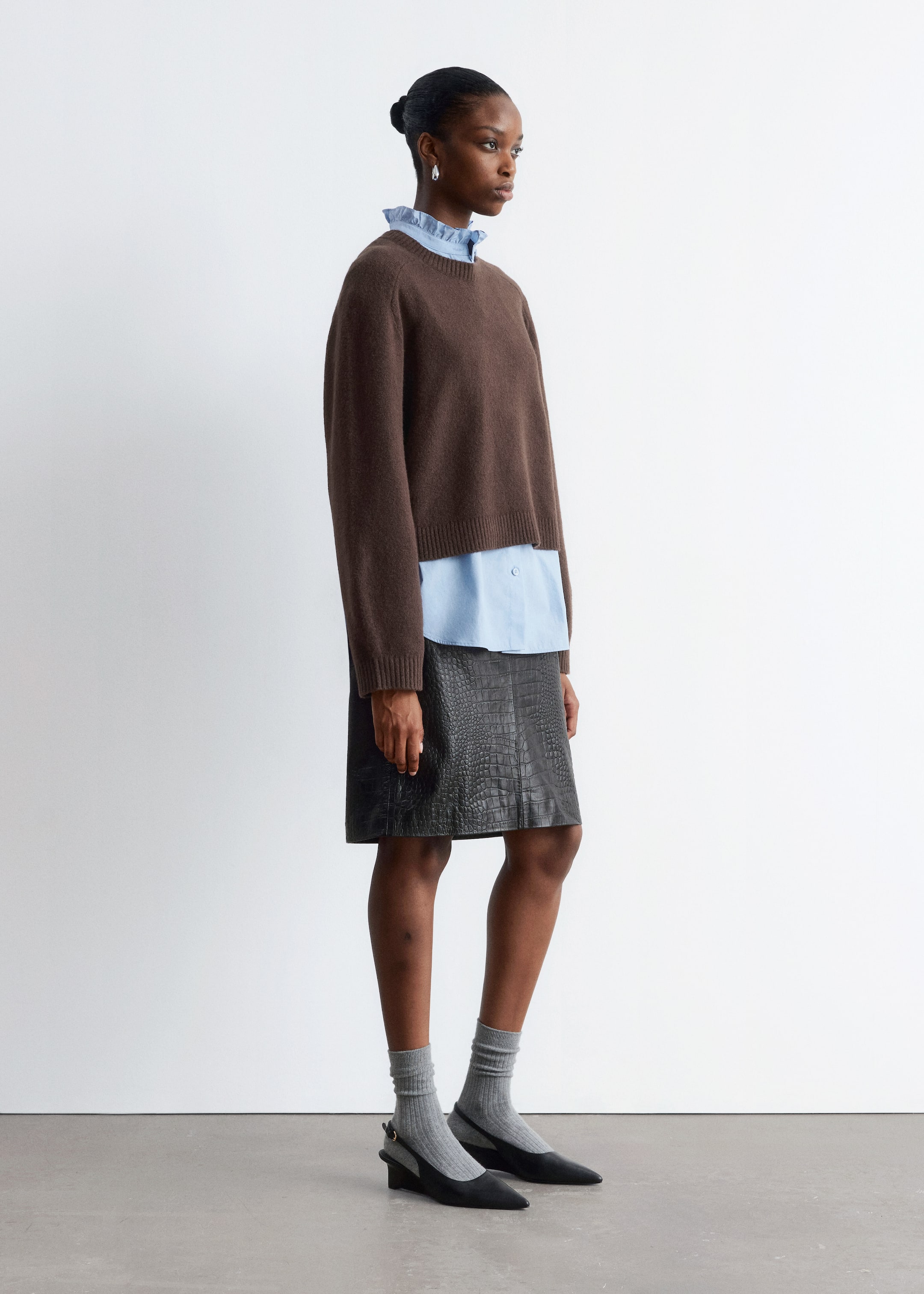 Grösseres Bild ansehen: Ein brauner Strickpullover mit Rundhalsausschnitt wird aus einem Seitenprofil gezeigt und hat eine entspannte, leicht verkürzte Passform. Er wird über einem hellblauen Hemd mit Kragen getragen, wobei der gerüschte Kragen und der Saum des Hemdes sichtbar unter dem Pullover hervorragen.