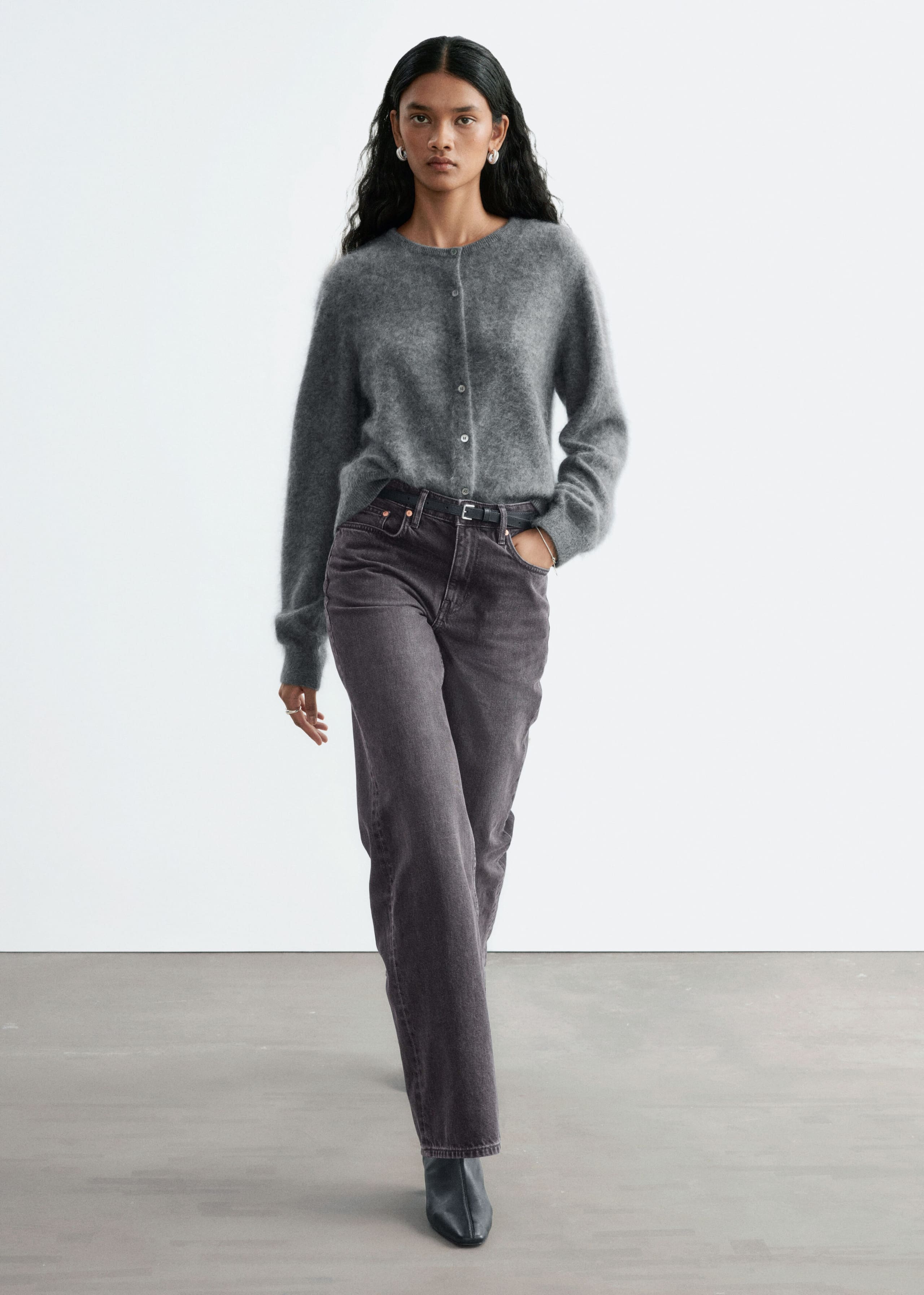 Straight-leg, mid-rise jeans in a faded dark gray wash are worn by the model, who is standing with one hand resting in a front pocket. The classic five-pocket jeans are styled with a thin black belt, a tucked-in gray cardigan, and black boots.