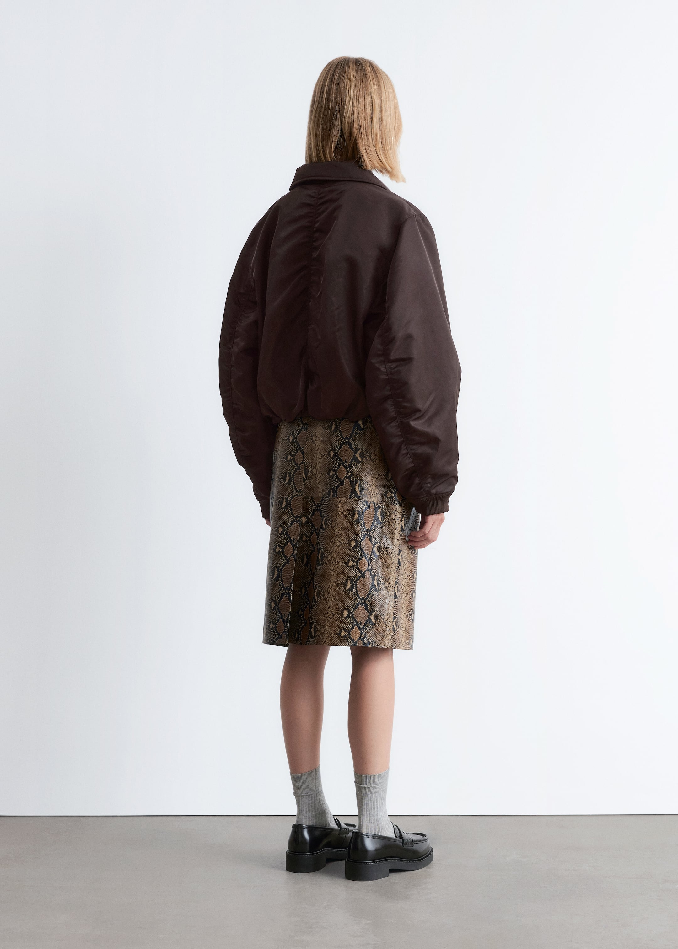 Ingrandisci l'immagine: Una giacca bomber marrone scuro è indossata, caratterizzata da un dettaglio arricciato sul retro e polsini e orlo elasticizzati. È abbinata a una gonna al ginocchio con stampa serpente marrone e nera, e mocassini neri sono indossati con calzini a coste grigio chiaro.