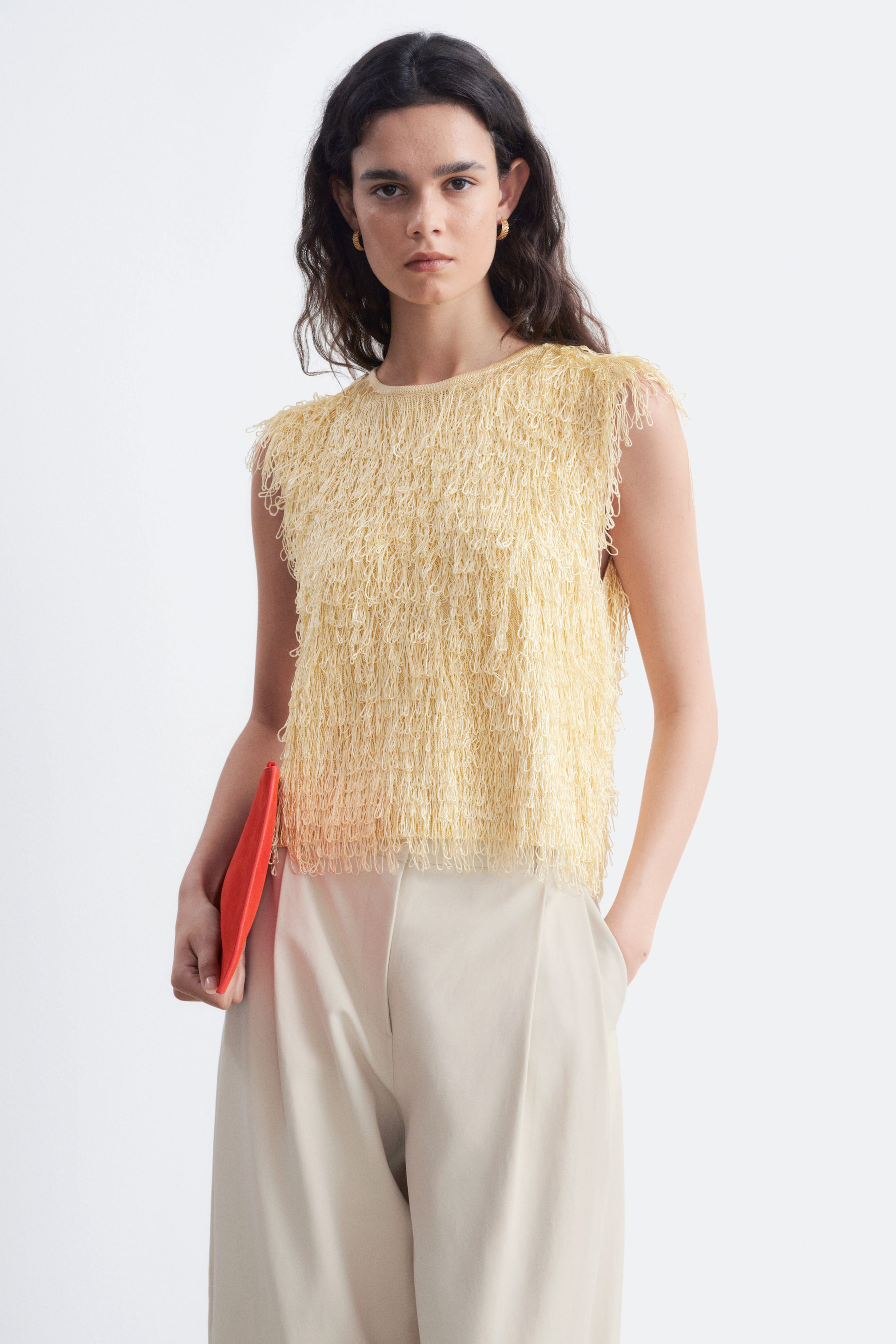 Visualizza l'immagine ingrandita: La modella indossa un top senza maniche di colore giallo chiaro. Presenta uno scollo rotondo ed è impreziosito da delicati frange annodate. La modella abbina il top a pantaloni beige chiaro, tenendo una pochette rossa in una mano mentre l’altra è appoggiata nella tasca dei pantaloni.