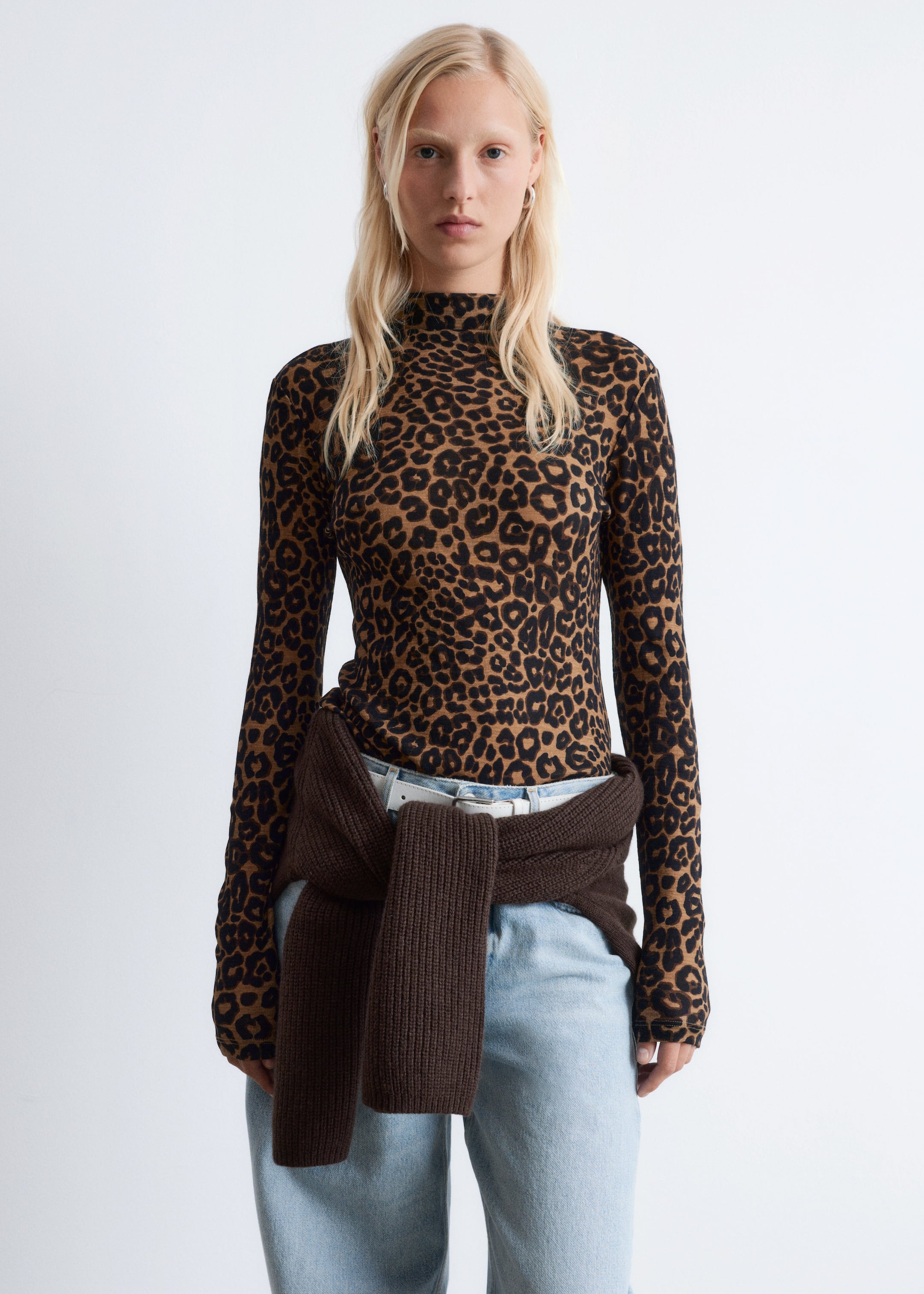 Visa större bild: En figursydd långärmad topp med ett brunt och svart leopardmönster och en hög hals visas framifrån, instoppad i ljusblå jeans med en brun stickad tröja knuten runt midjan.