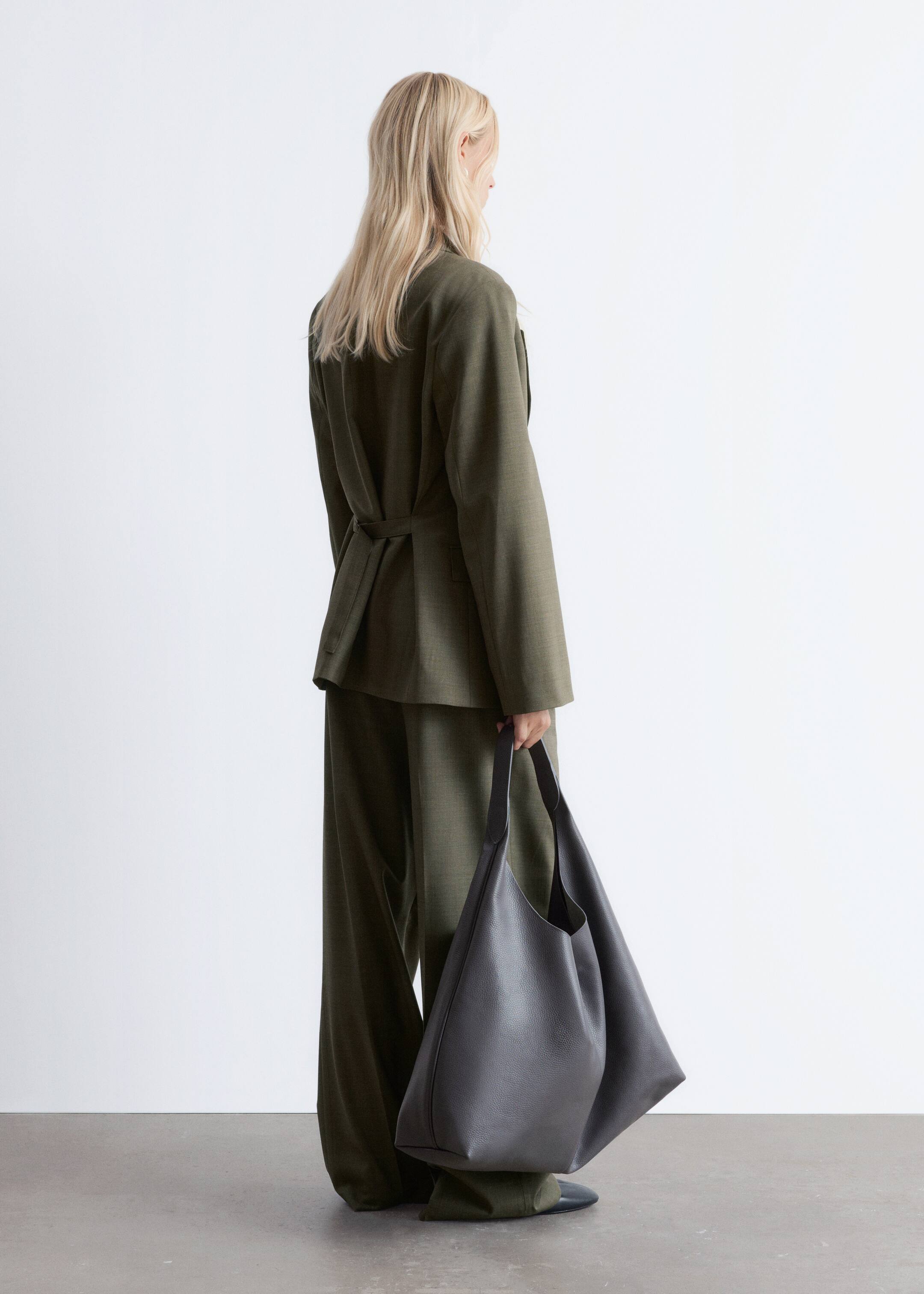 Visualizza immagine più grande: Un blazer verde oliva scuro e pantaloni coordinati a gamba larga sono presentati di dorso, mostrando il dettaglio del blazer con cintura in vita e il fit rilassato di entrambi i capi. Una grande borsa tote in pelle scura grigia ciottolata e non strutturata viene portata nella mano destra.