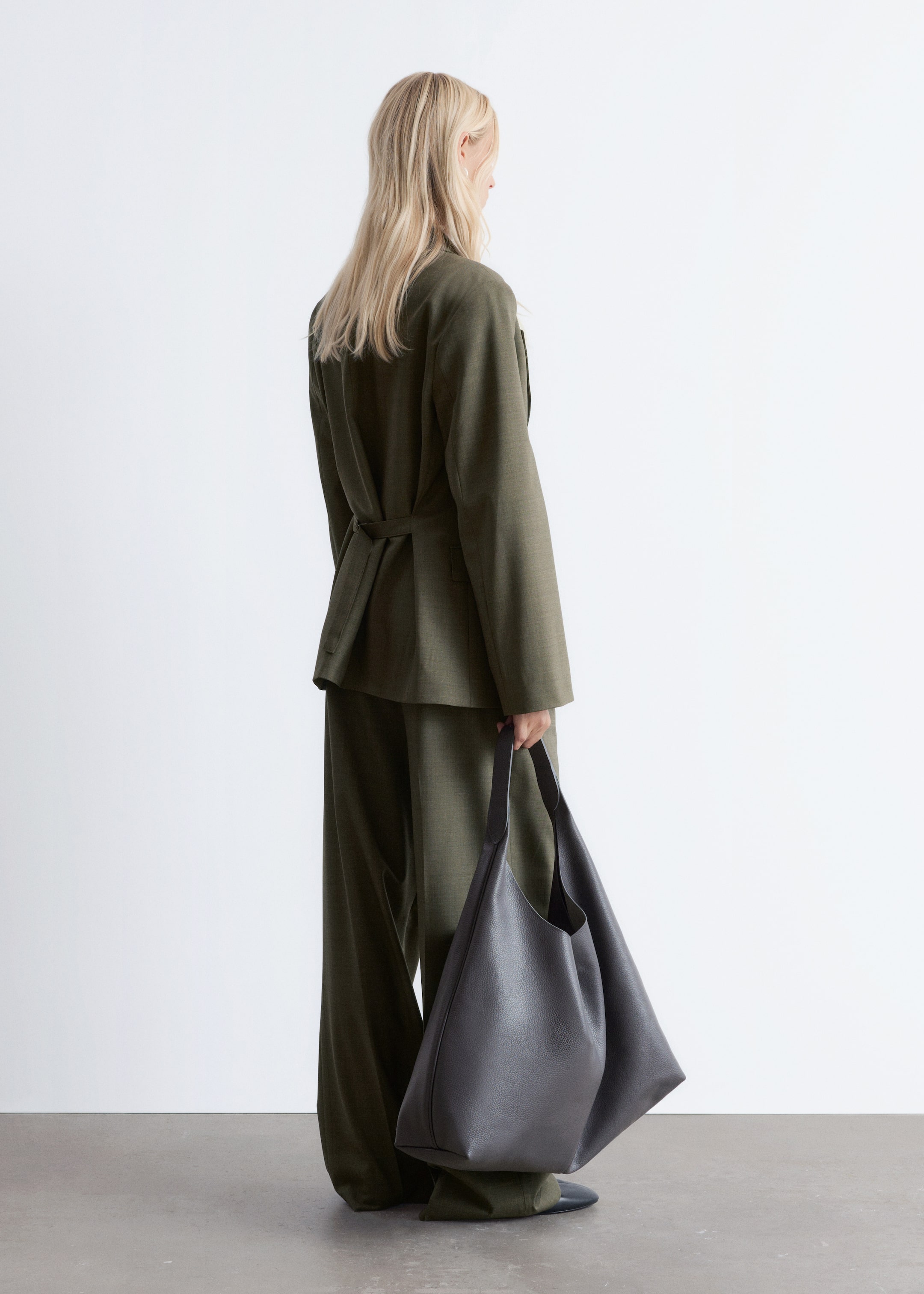 Ingrandisci l'immagine: Un blazer verde oliva scuro e pantaloni abbinati a gamba larga sono presentati dal retro, mostrando il dettaglio del blazer con cintura in vita e la vestibilità rilassata di entrambi i capi. Una grande borsa tote in pelle scura grigia non strutturata e con effetto granuloso è portata nella mano destra.