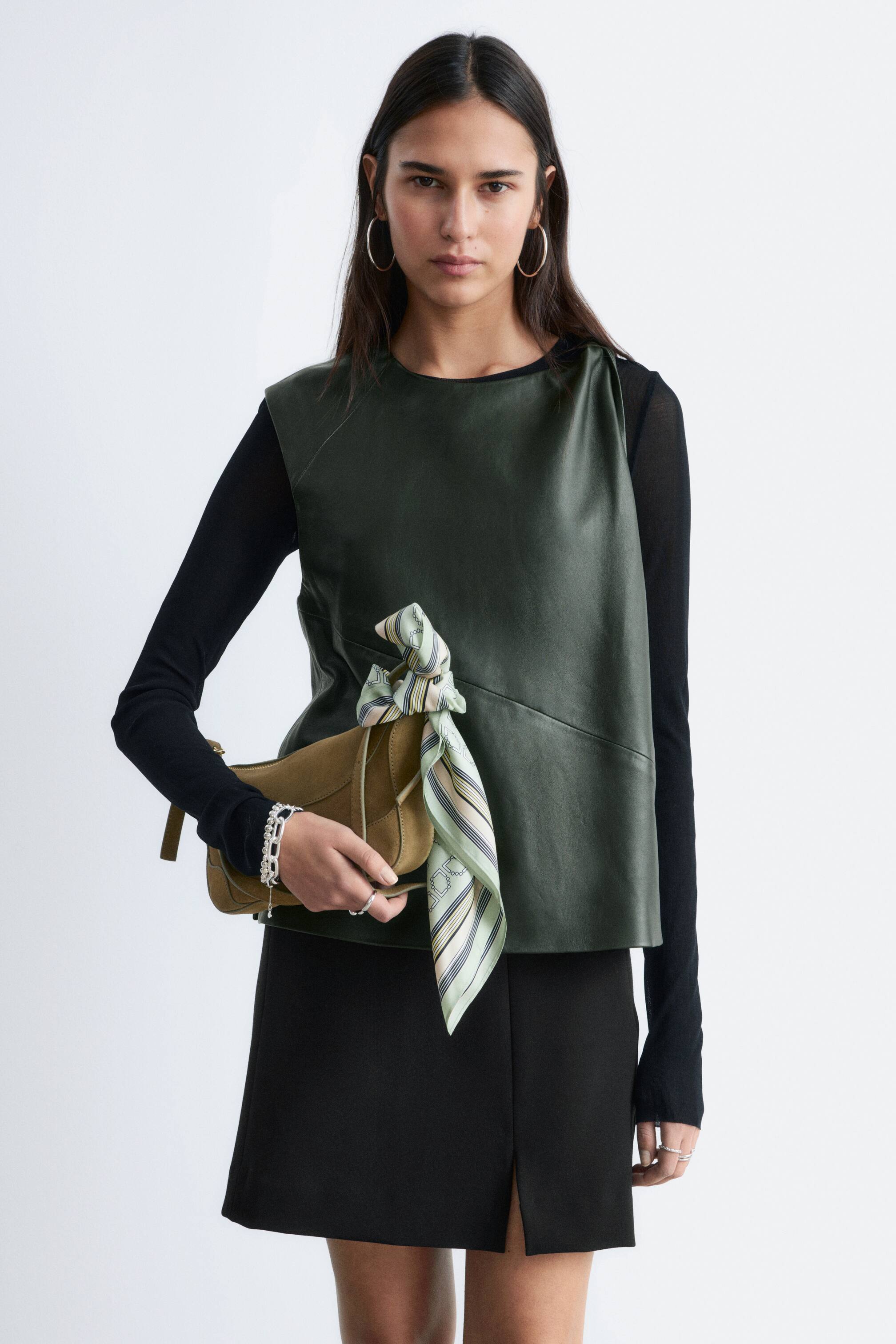 Visualizza immagine più grande: Una borsa a tracolla in pelle opaca verde oliva viene tenuta, leggermente inclinata verso destra, mettendo in risalto una chiusura a patta curva. Un foulard a righe verde chiaro, beige e rosa è annodato intorno al suo cinturino, caratterizzato da un motivo geometrico.