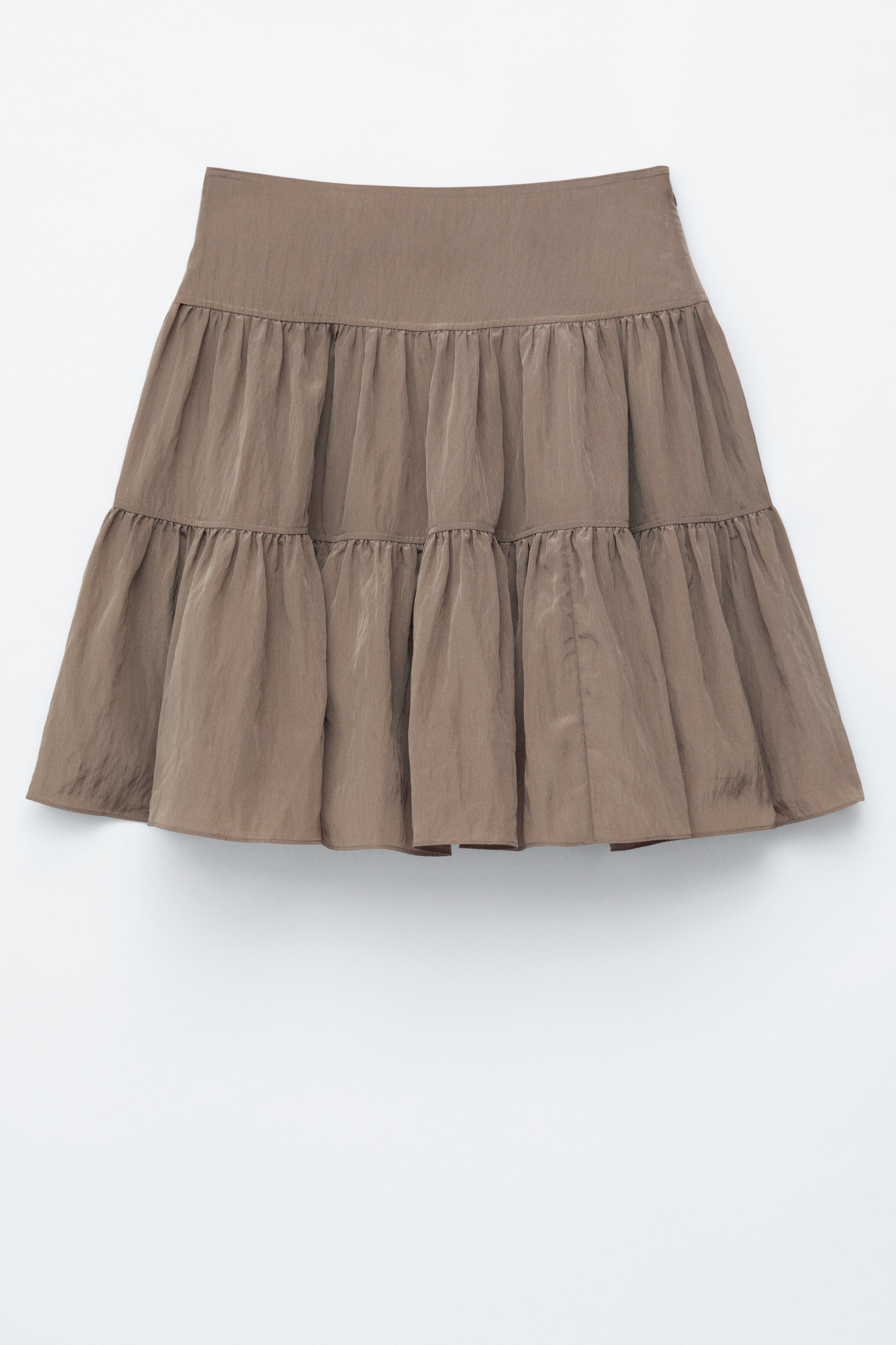 Vis større bilde: En taupefarget lagdelt skjørt vises fra forsiden, med et flatt linning og to rynkede volanglag. Stoffet har en subtil glans og en krøllete tekstur.