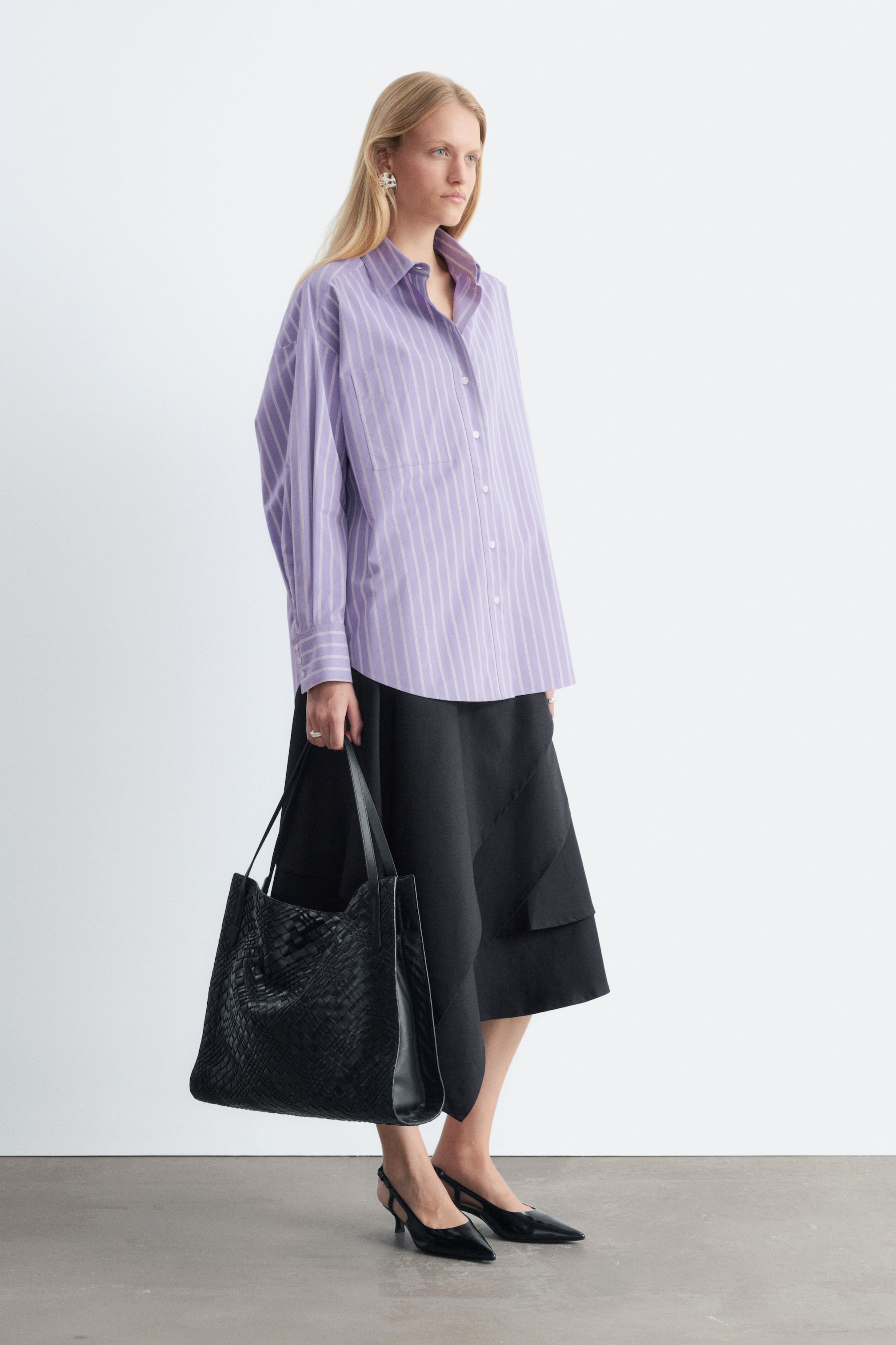 Agrandir l'image: Le modèle, debout de profil, porte une chemise oversize à rayures verticales lilas et blanches. La chemise présente un col classique, des manches longues et une poche plaquée sur la poitrine, et elle est portée non rentrée sur une jupe midi noire, tandis que le modèle tient un cabas tressé noir.