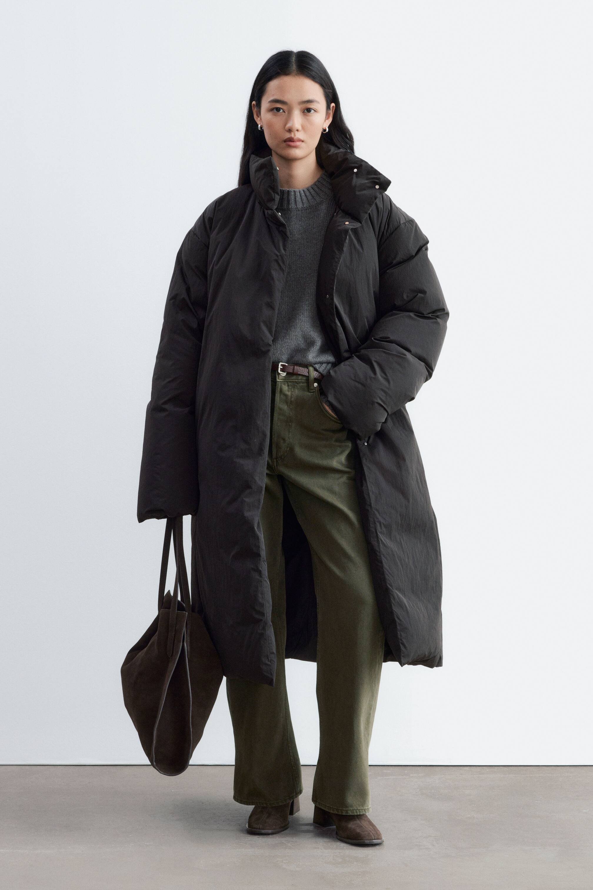 View larger image: A long black down jacket, featuring a loose silhouette and a high funnel collar, is being worn open by the model. The jacket is layered over a grey crew-neck sweater and olive-green trousers, with a dark brown suede tote bag being carried in one hand.