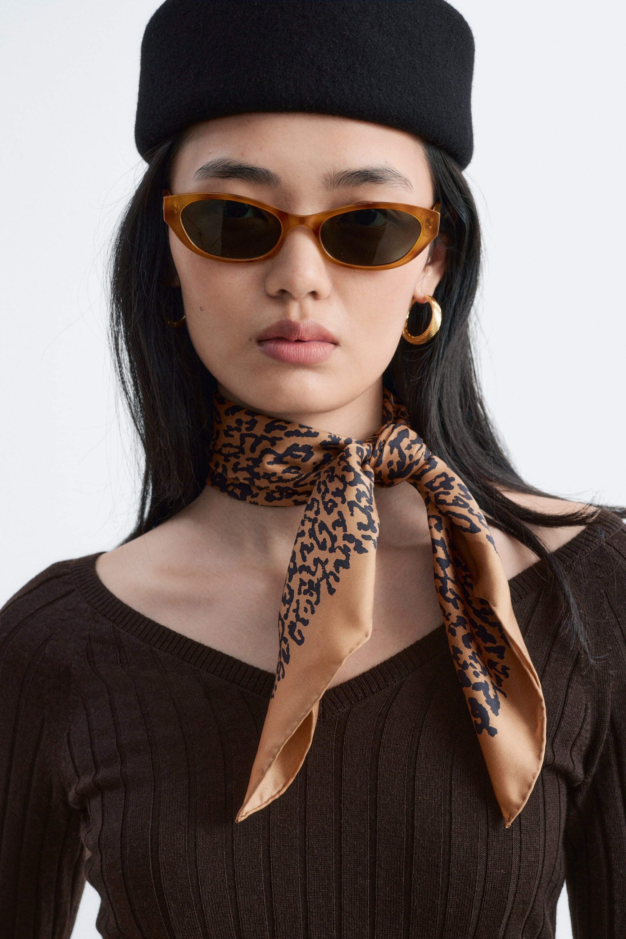 Zobraziť väčší obrázok: Modelka má na sebe tmavohnedý rebrovaný vlnený top slim strihu s výstrihom do V, doplnený hodvábnou šatkou s leopardím vzorom uviazanou okolo krku. Modelka má tiež čierny klobúk a hnedé slnečné okuliare.