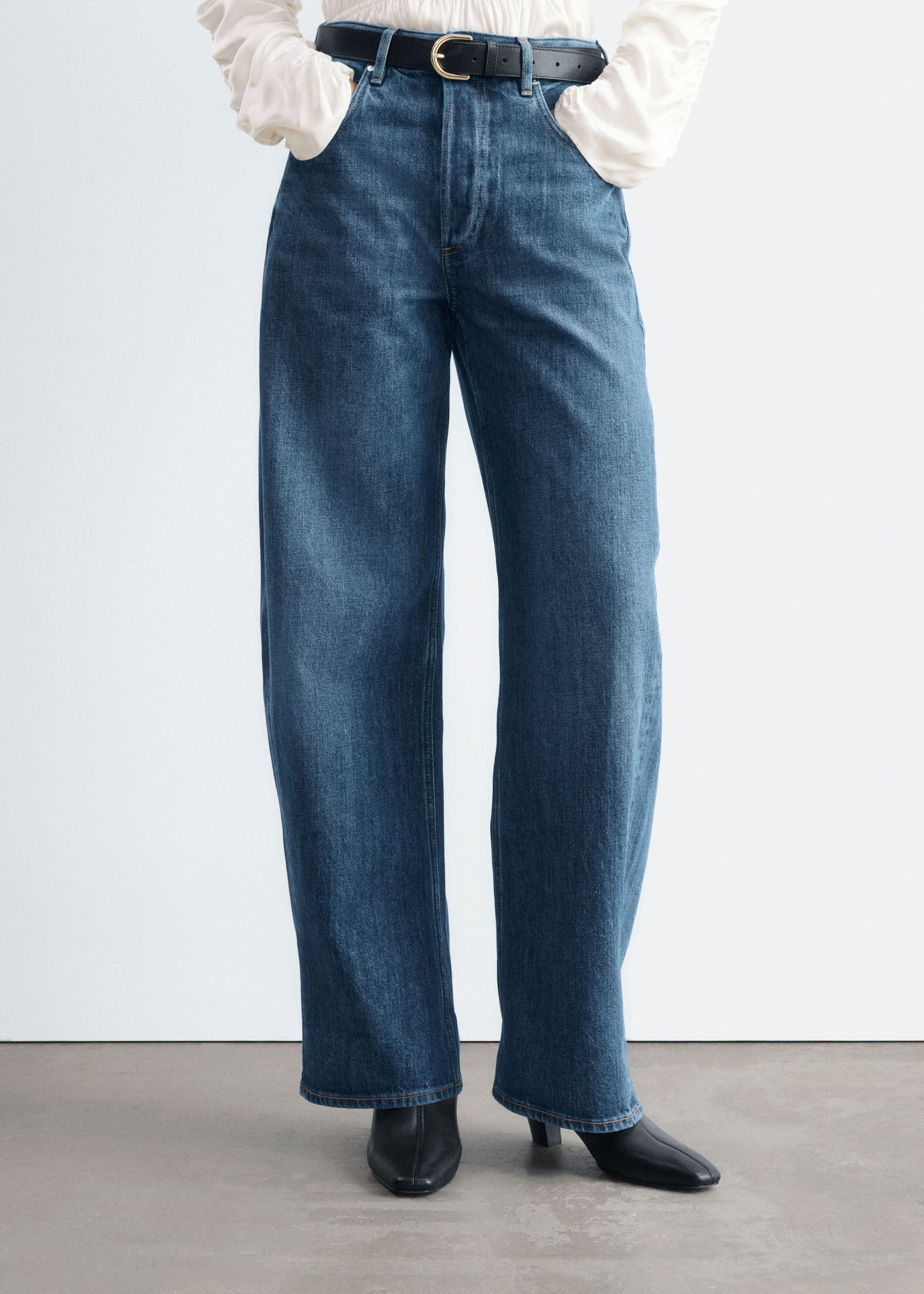Blue denim barrel-leg jeans with an oversized fit are worn by the model, who is standing with her hands in the front pockets. The jeans are styled with a black leather belt, a tucked-in white blouse, and black heeled boots.