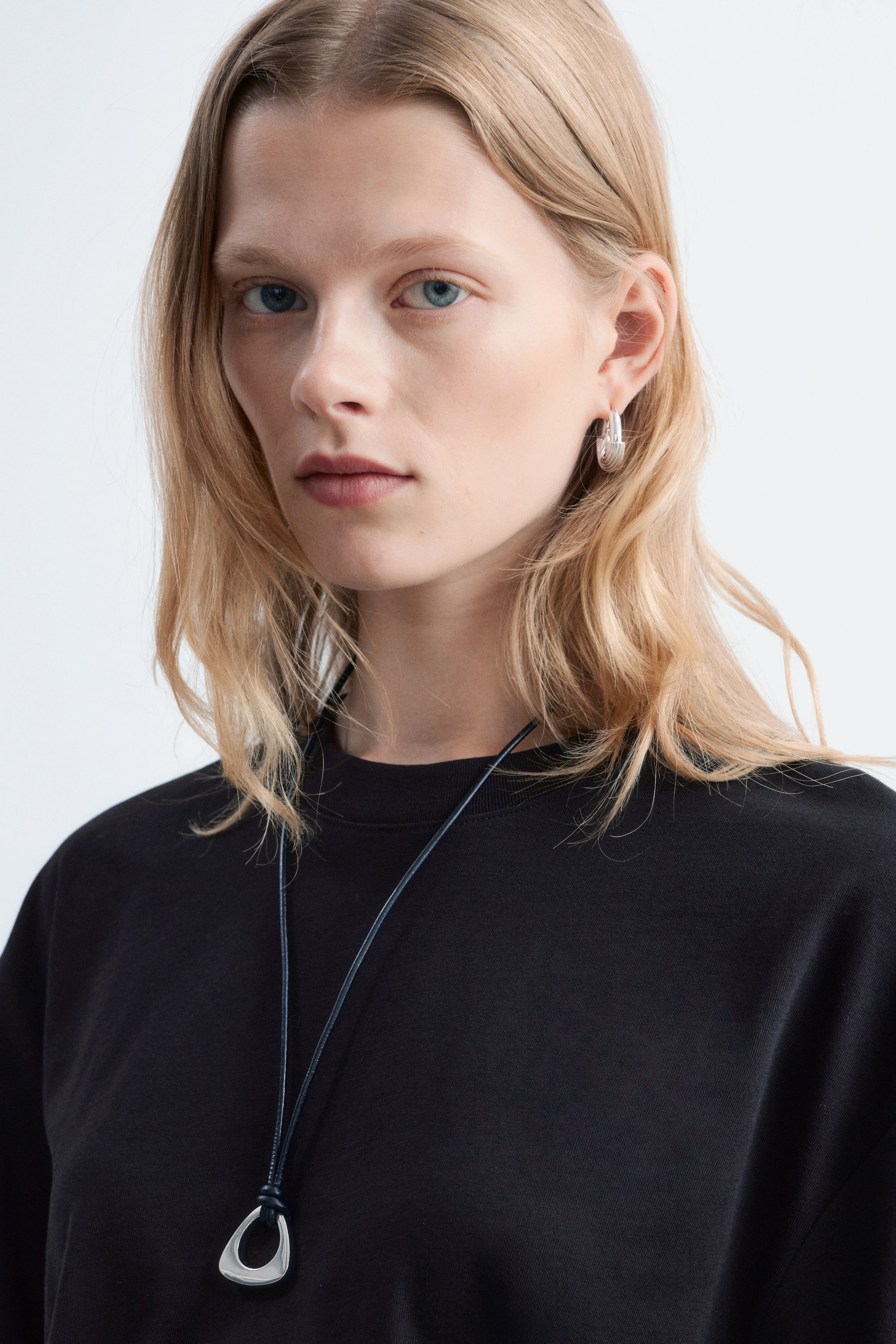 Grotere afbeelding bekijken: Een zwart T-shirt met een ronde hals en een ontspannen pasvorm wordt gedragen door het model. Een donker koordketting met een zilverkleurige hanger rust op het T-shirt.