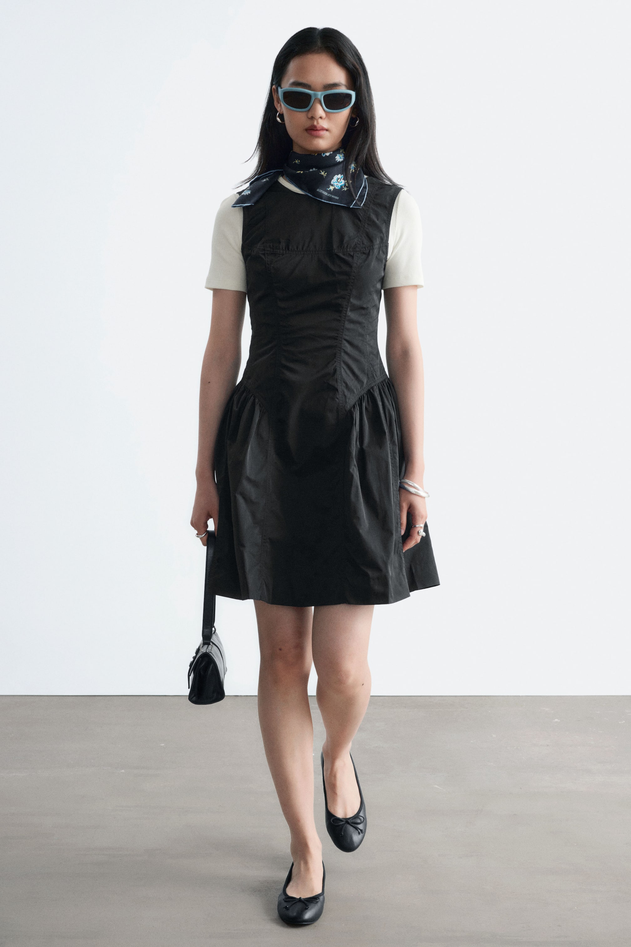 Ver imagen más grande: Una minivestido peplum negro con corsé estructurado y falda fruncida y acampanada lo lleva la modelo, superpuesto sobre una camiseta de manga corta color crema. El vestido se combina con un pañuelo estampado al cuello, gafas de sol azul claro, bailarinas negras y un pequeño bolso negro que lleva en una mano.