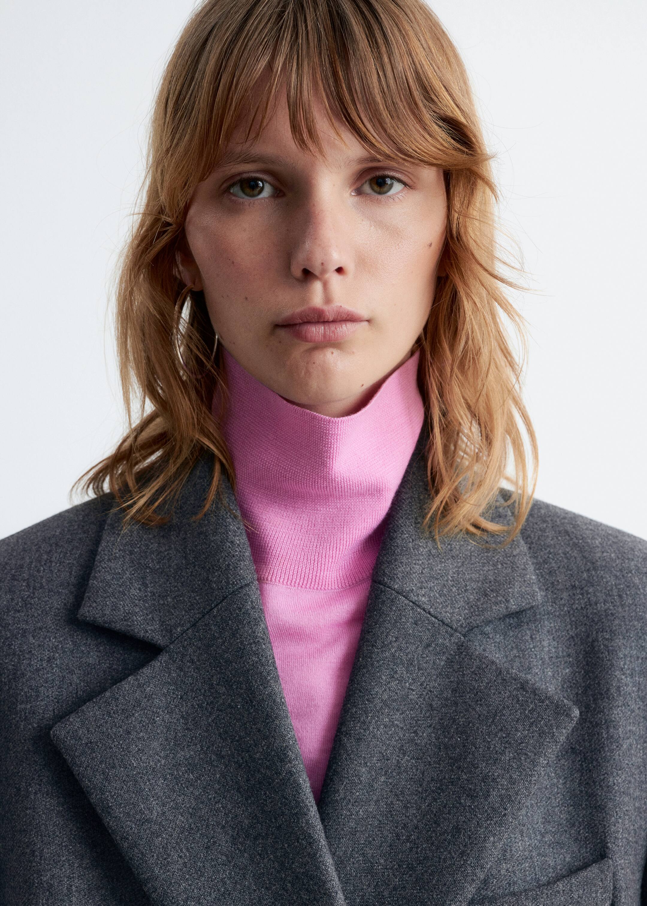 Grösseres Bild ansehen: Ein dunkelgrauer Blazer mit Kerbrevers wird über einem leuchtend pinken Rollkragenpullover mit geripptem Kragen getragen.