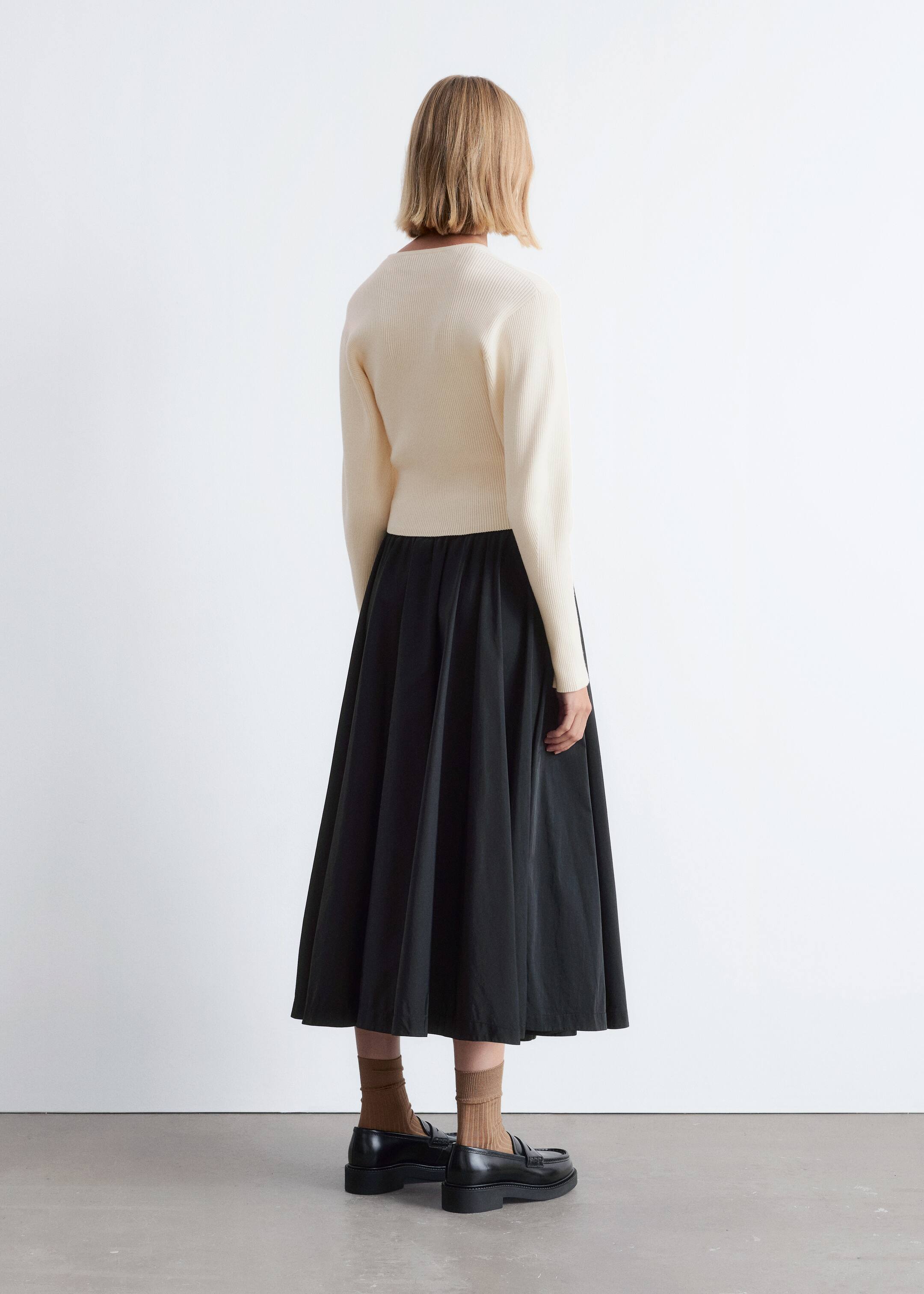 Größeres Bild anzeigen: Ein cremefarbener gerippter Stricktop, der von hinten sichtbar ist, wird über einem schwarzen, vollen Midirock mit geraffter Taille getragen, der bis zu den Waden fließt. Schwarze Loafer mit dicken Sohlen werden mit braunen gerippten Socken getragen.