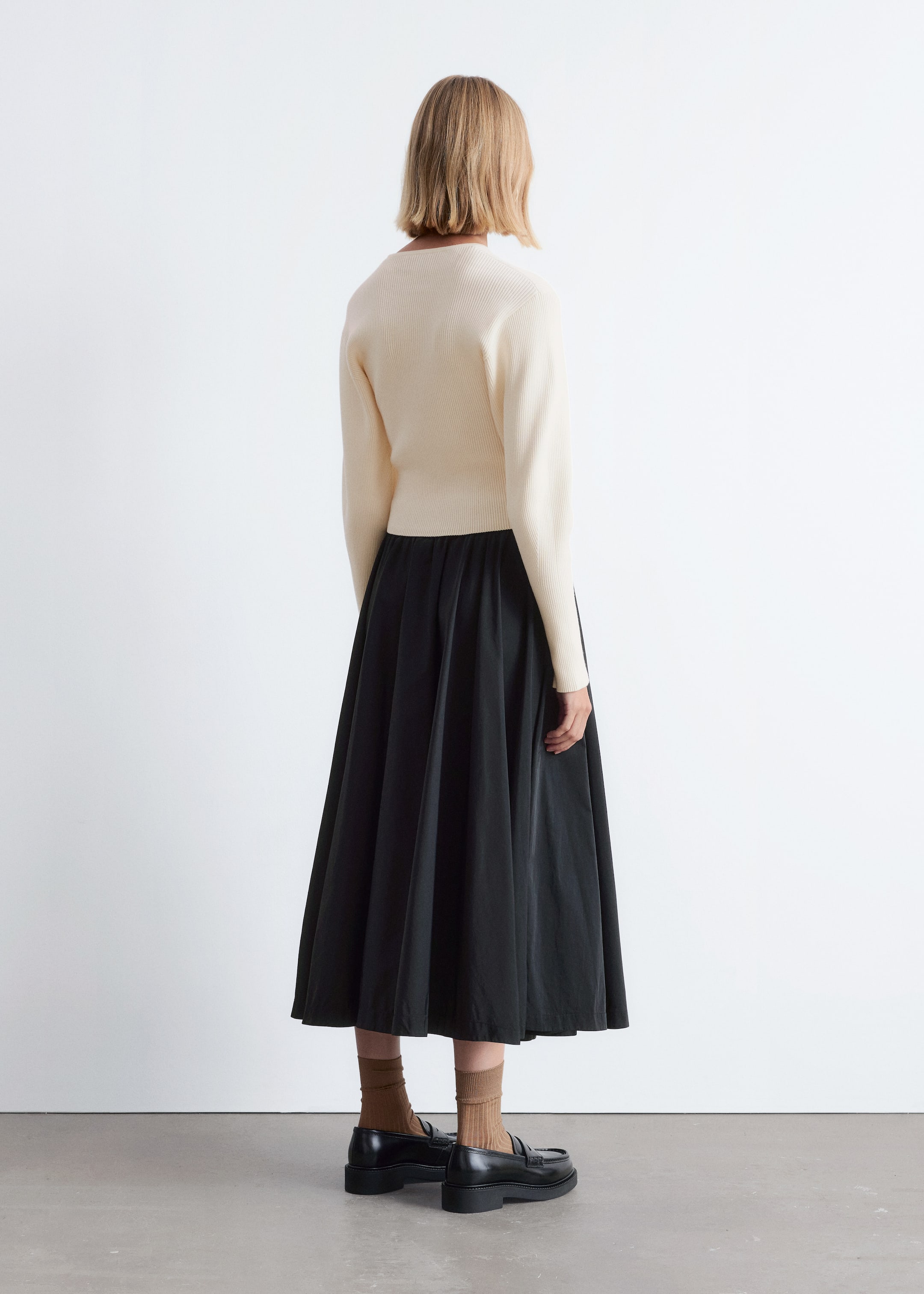 Grösseres Bild ansehen: Ein cremefarbener gerippter Stricktop, der von hinten sichtbar ist, wird über einem schwarzen, vollen Midirock mit geraffter Taille getragen, der bis zu den Waden fließt. Schwarze Loafer mit dicken Sohlen werden mit braunen gerippten Socken getragen.