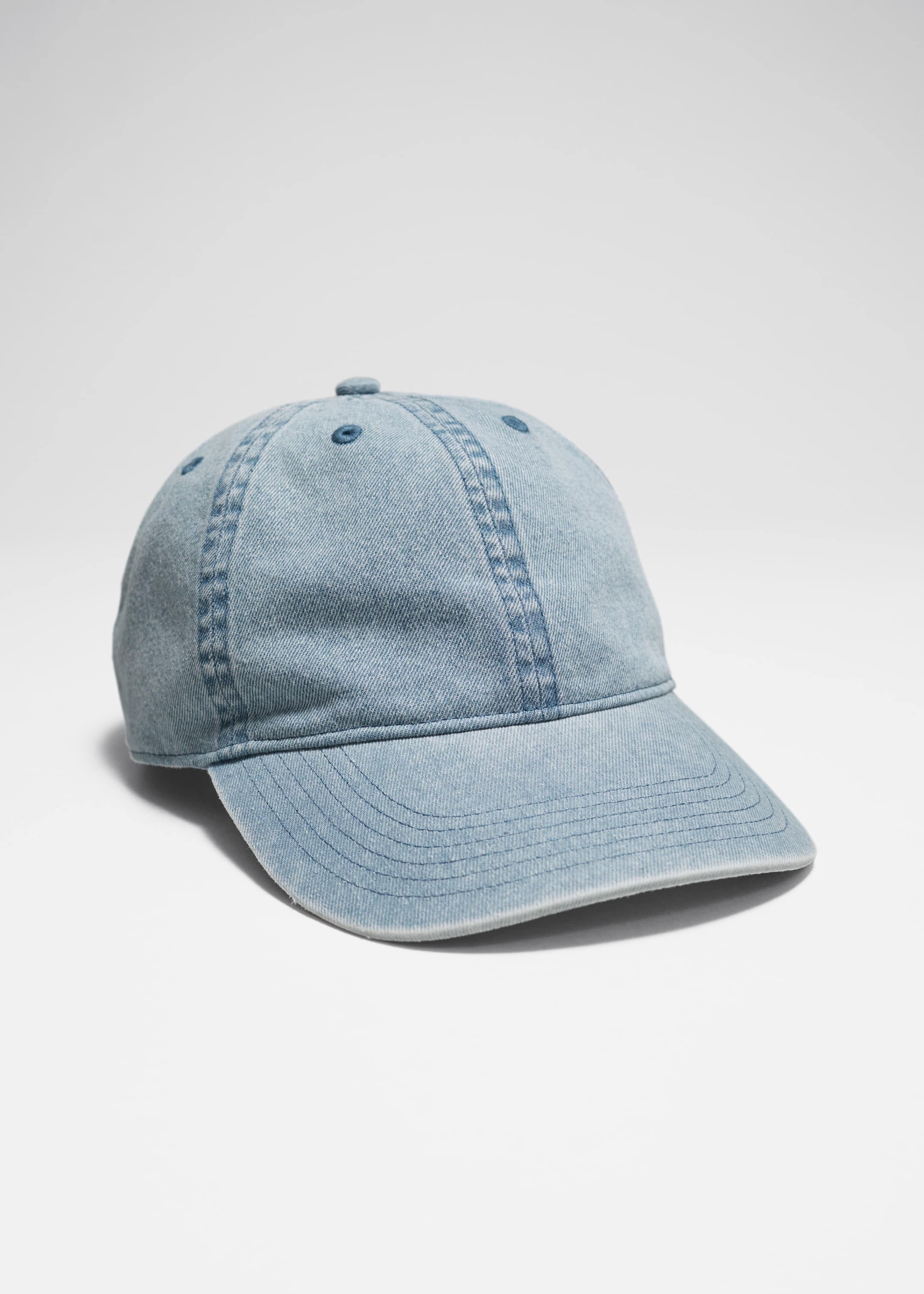 Visualizza immagine più grande: Un berretto da baseball in denim lavato azzurro è inclinato leggermente verso destra. La sua corona a sei pannelli è dettagliata con cuciture a vista e piccoli occhielli, e la sua visiera curva mostra più linee di cucitura parallele.