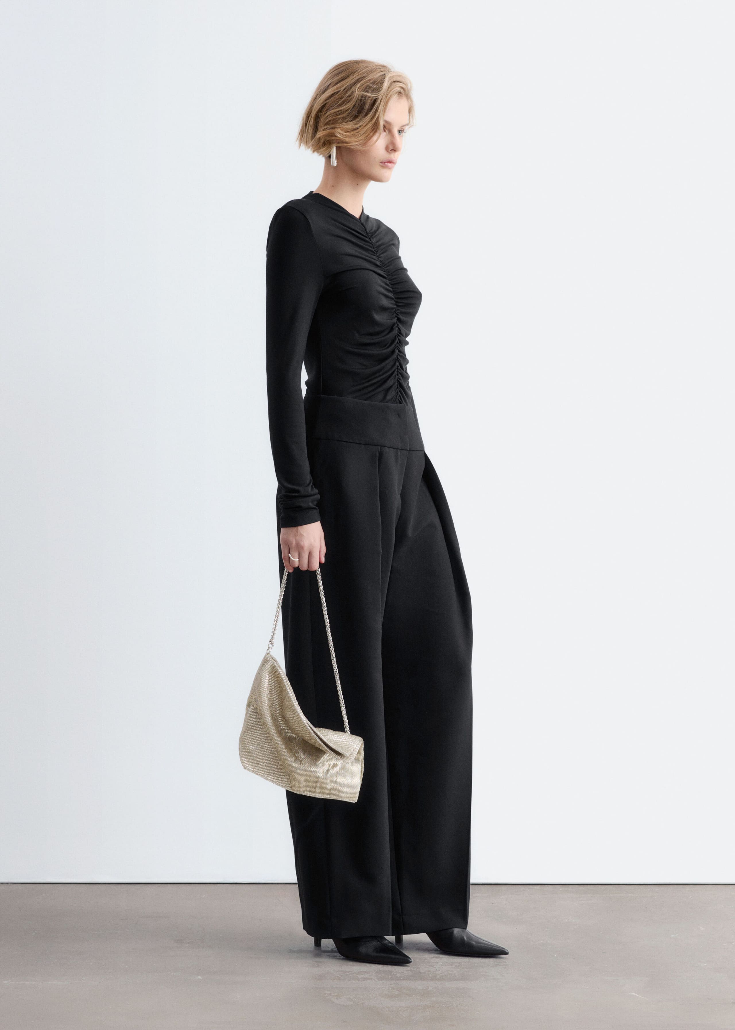 A black, long-sleeved top with gathered ruching along the center-front seam is worn by the model. The fitted top is tucked into wide-leg black trousers and styled with a metallic shoulder bag held in one hand.