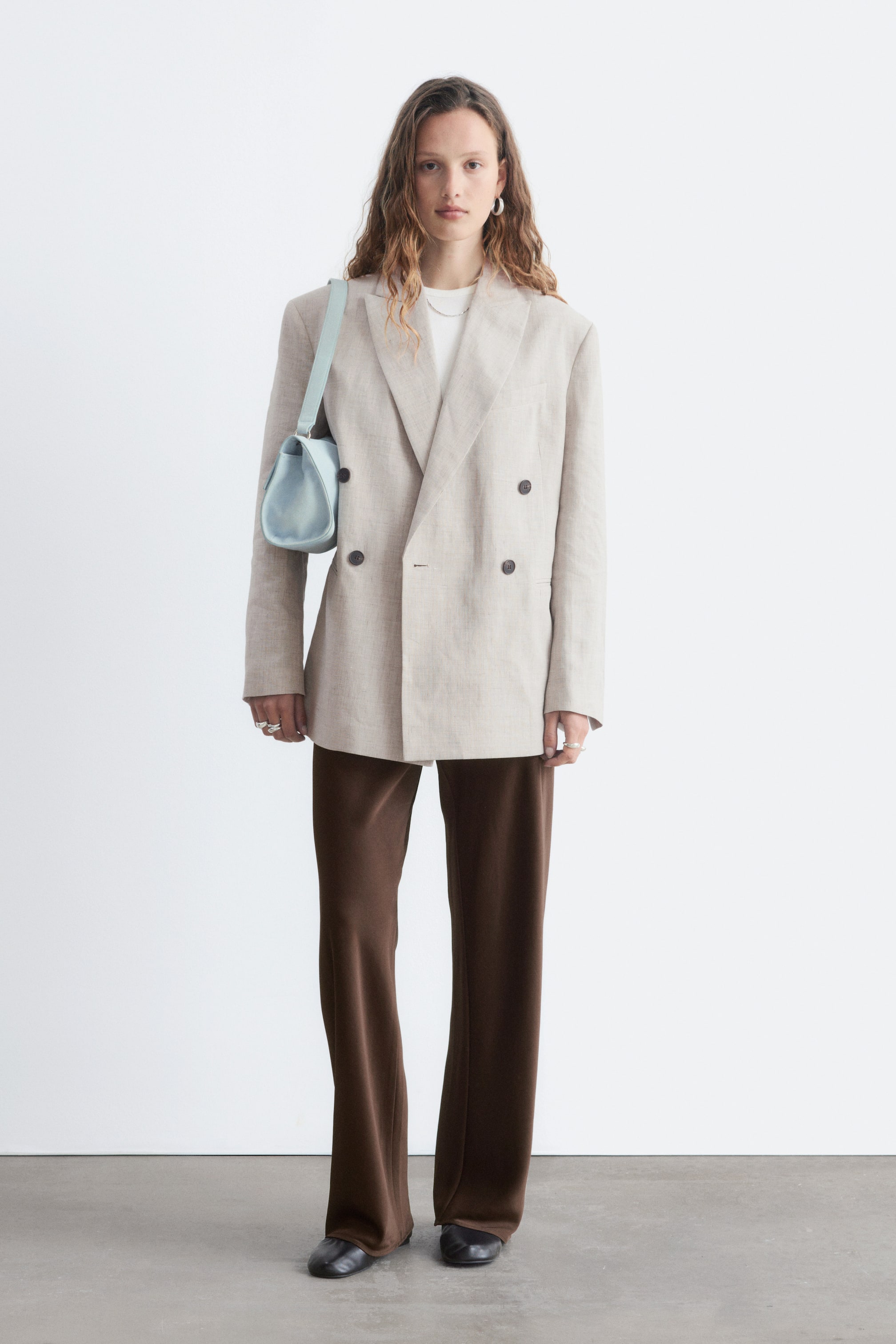 Vis større bilde: En beige dobbeltspent blazer med en oversized passform, strukturerte skuldre, hakket jakkeslag og fire mørke knapper foran. Den er stylet åpen over en hvit t-skjorte, med en lyseblå skulderveske hvilende på venstre side.