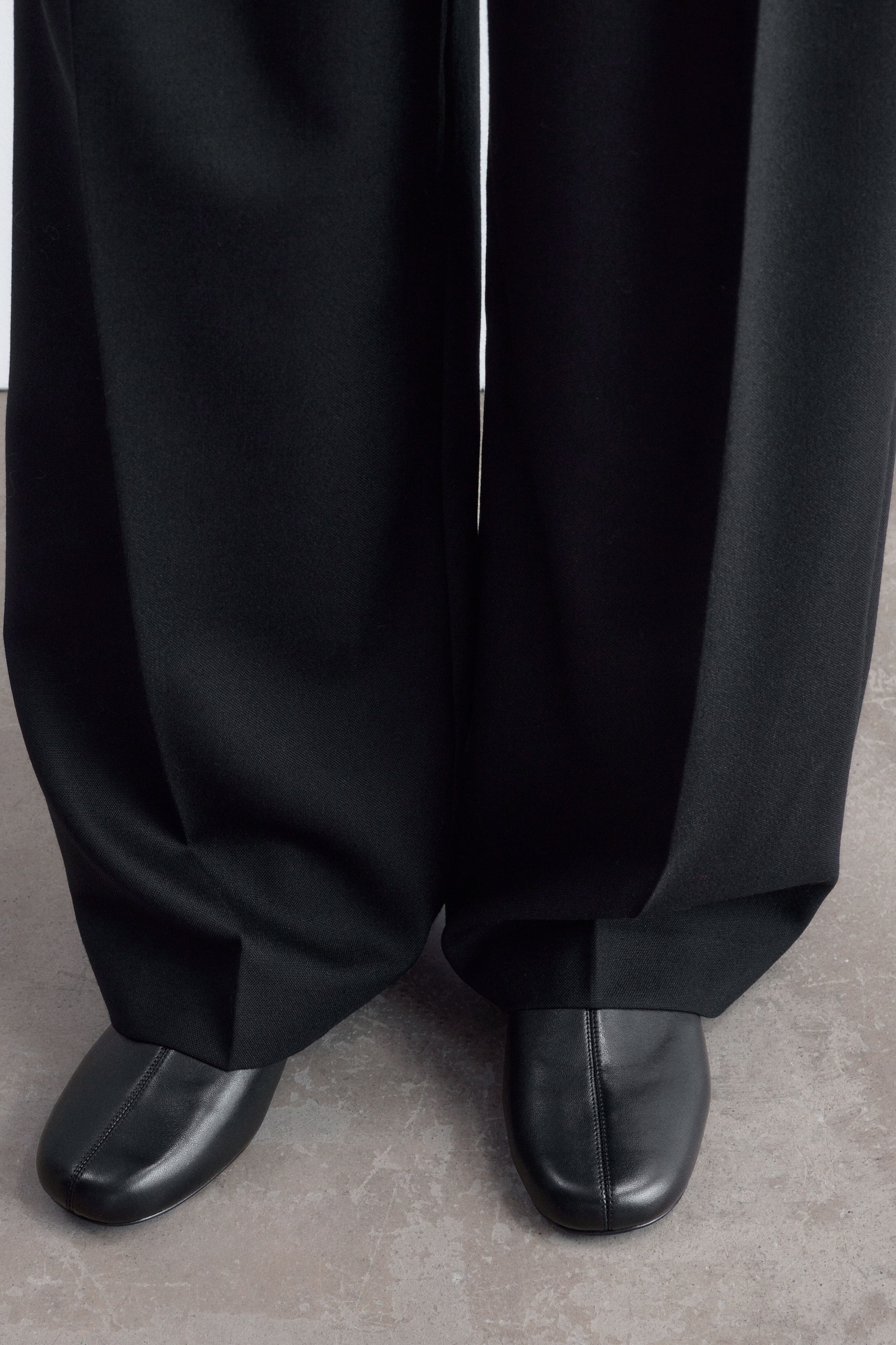 View larger image: Black leather boots featuring a rounded toe and central seam are worn by the model, with wide-leg black trousers draping over them.