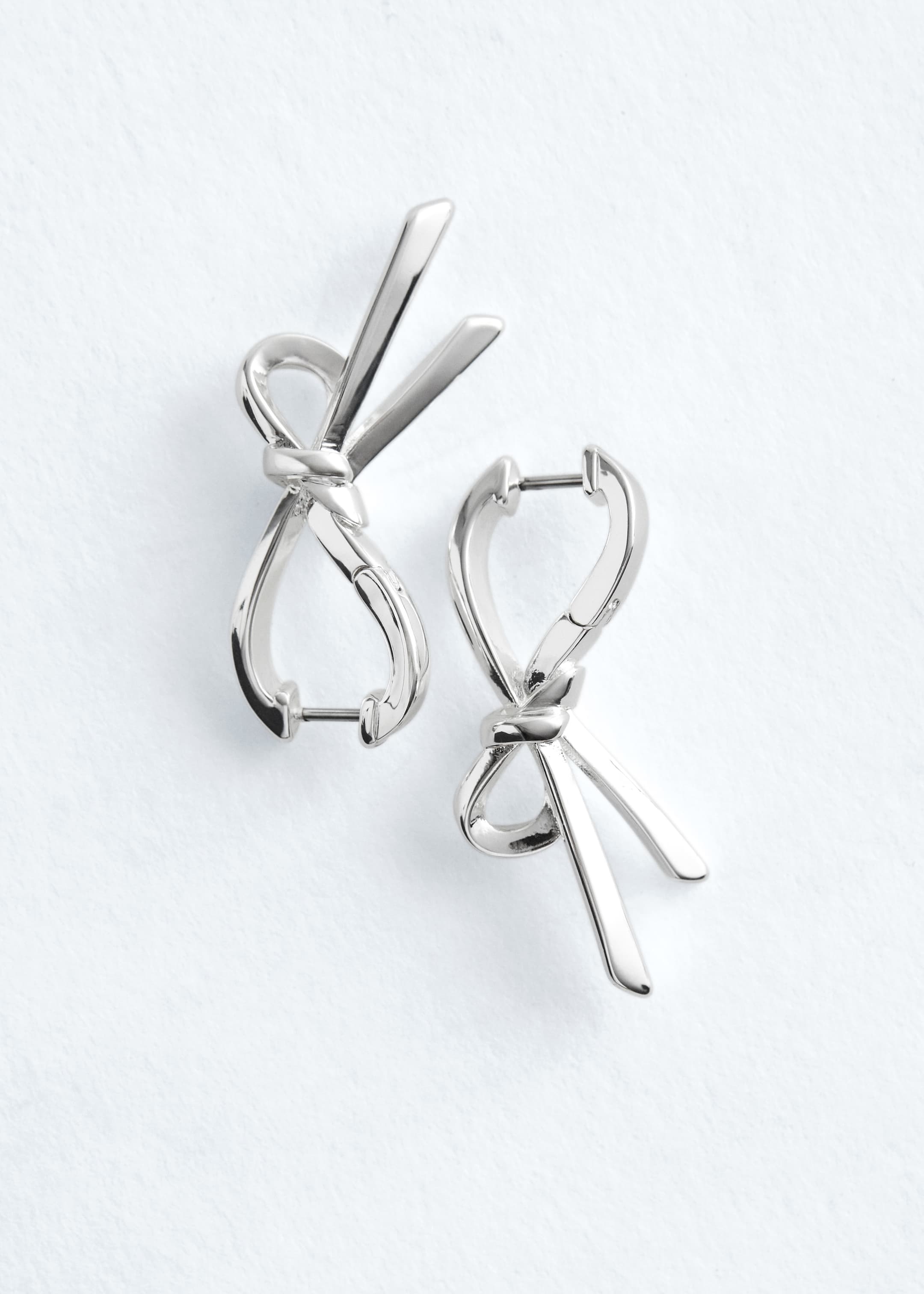 Ver imagem maior: Um par de brincos de argola em tom prateado exibe um design de laço estilizado. Cada brinco é formado por metal liso e brilhante, apresentando dois laços e dois elementos pendentes em forma de fita, com fechos articulados no topo.