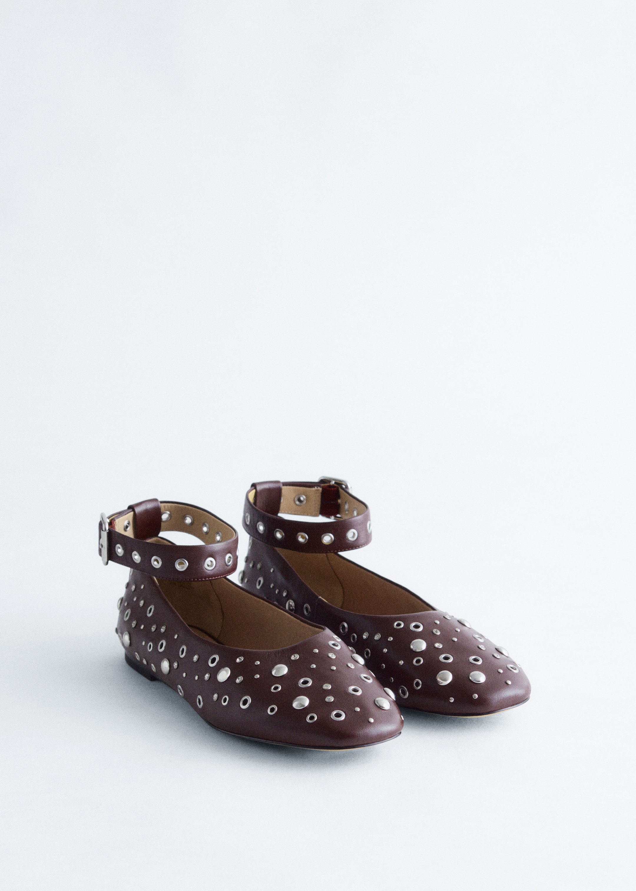 Grotere afbeelding bekijken: Een paar donkerrode leren ballerina's wordt gepresenteerd, iets schuin van voren. De schoenen zijn versierd met talrijke verspreide platte zilverkleurige studs en oogjes over de neus en zijkanten, en brede, bijpassende leren enkelbanden met zilverkleurige gespen en oogjes zijn bevestigd en staan rechtop.