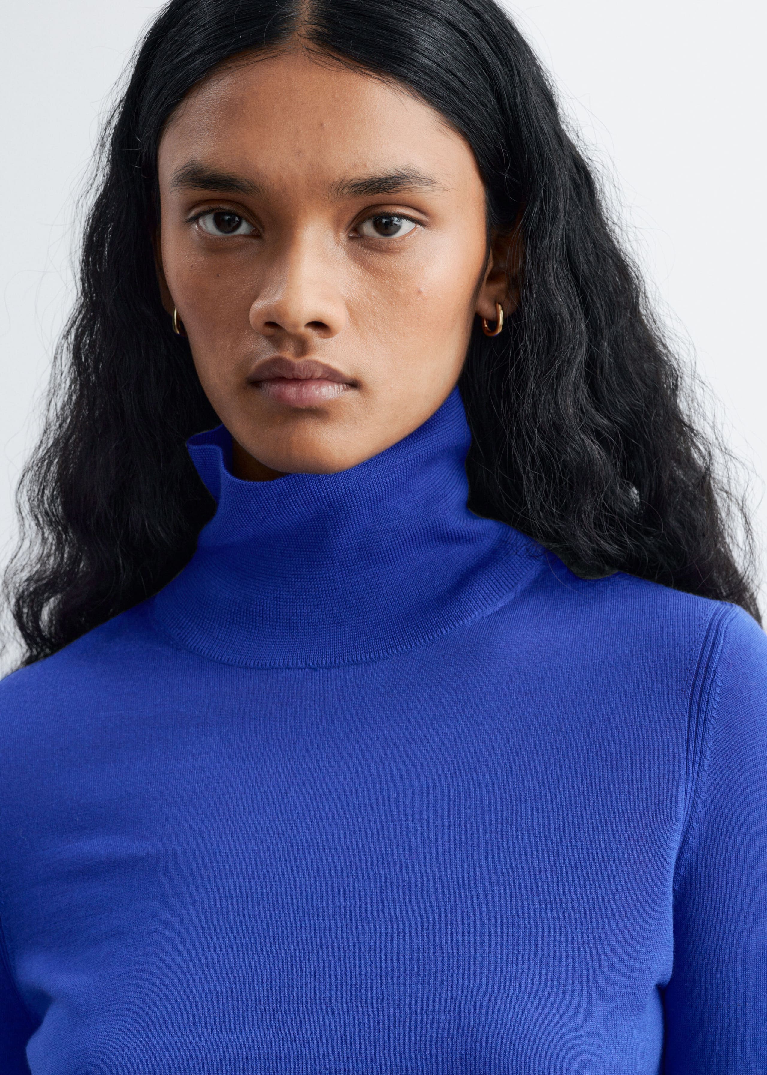 Een felblauwe polo met col van merinowol, voorzien van een hoge geribde kraag en een fijne gebreide textuur, wordt gedragen door het model. De close-up benadrukt de gestructureerde halslijn en de zachte stof van het kledingstuk.