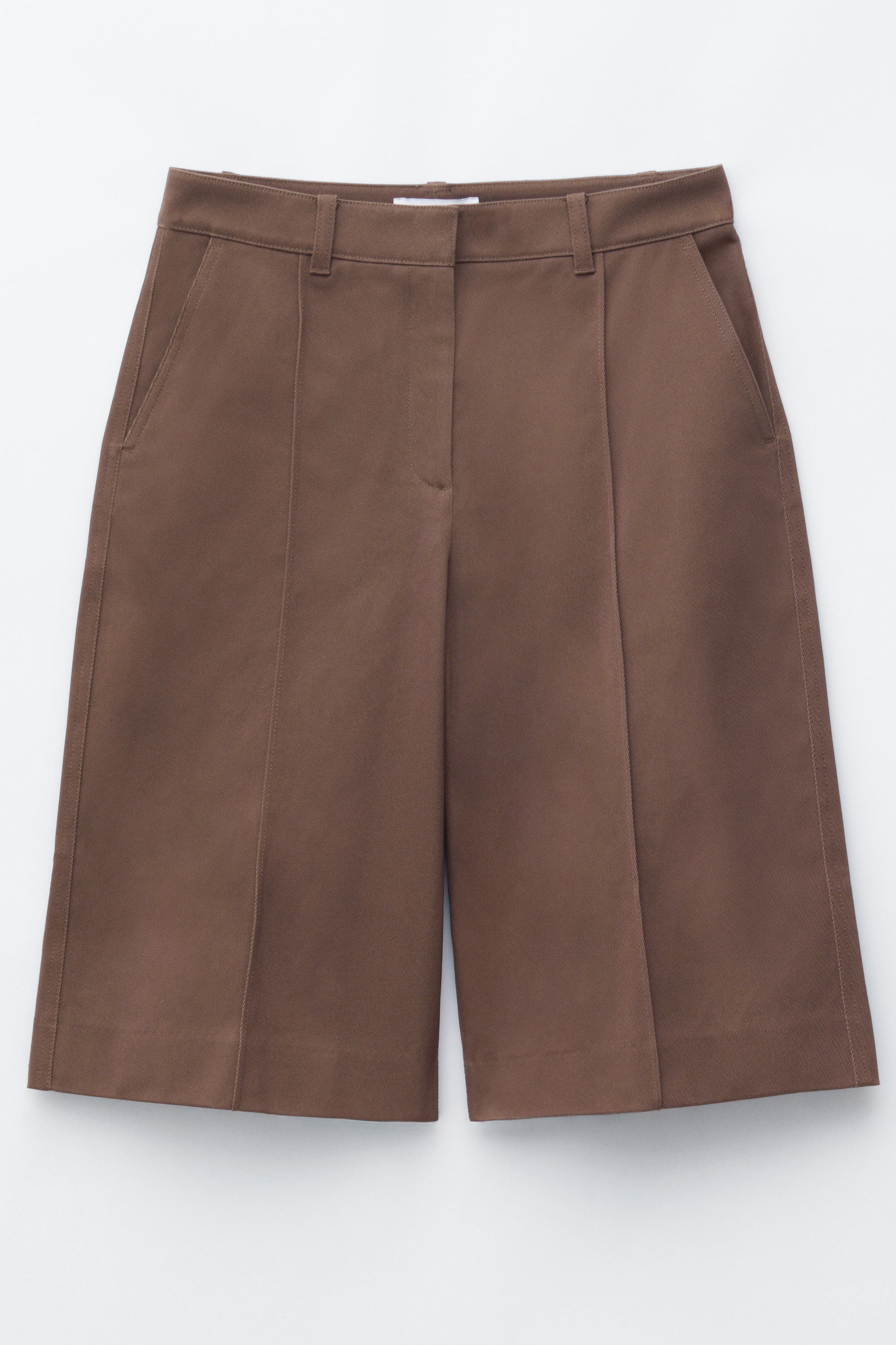 Zobrazit větší obrázek: Hnědé bermudské šortky jsou zobrazeny zepředu, s páskem s poutky na opasek, šikmými bočními kapsami a zipem s háčkem a očkem. Svislé švy s horním prošitím zdůrazňují přední část každé volně střižené nohavice, která sahá ke kolenům.
