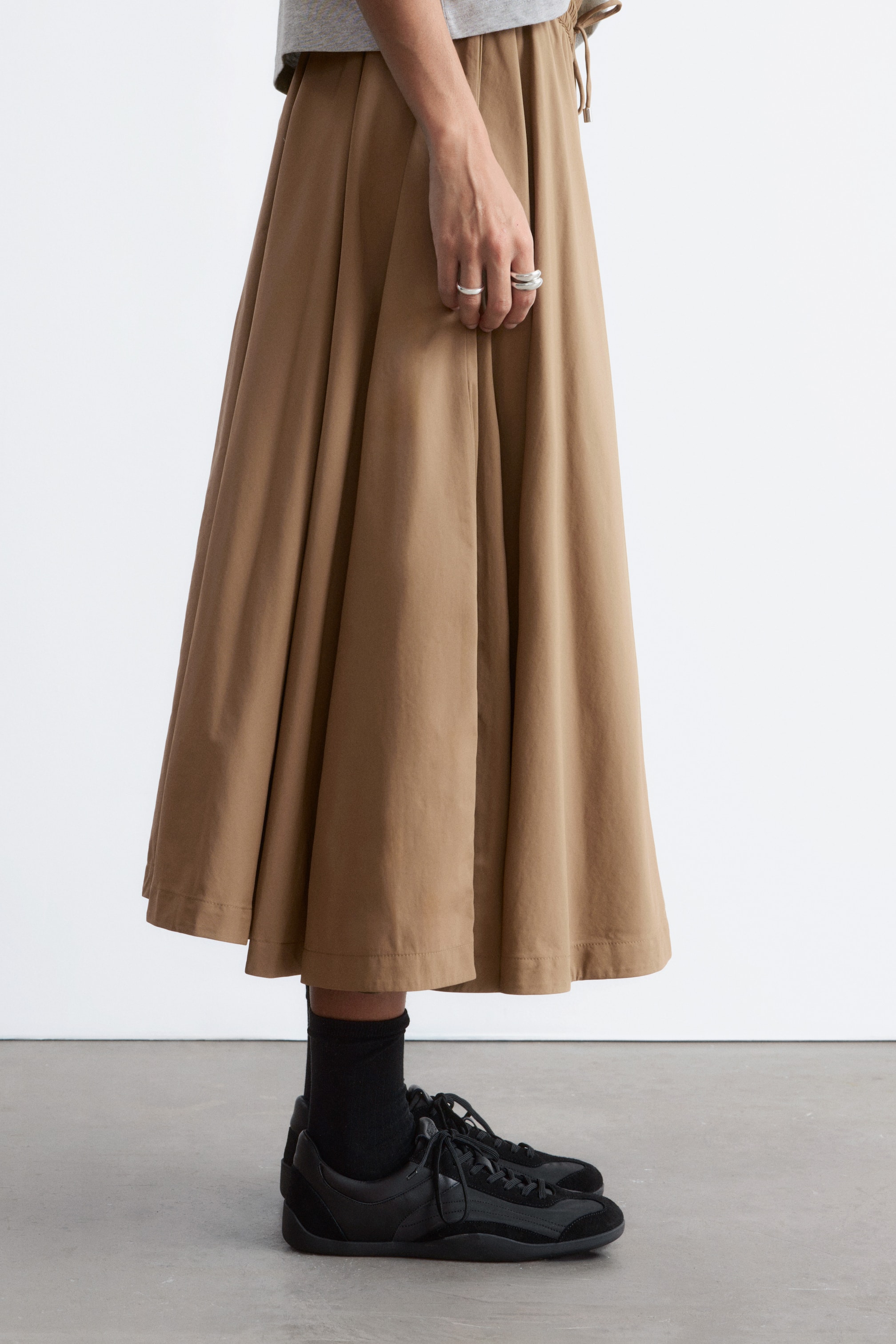 Größeres Bild ansehen: Ein beige Midirock mit einer vollen, A-Linien-Silhouette, sichtbaren Raffungen und einem Kordelzug in der Taille. Er fällt anmutig bis knapp über die Knöchel und präsentiert sein Volumen aus der Seitenansicht.