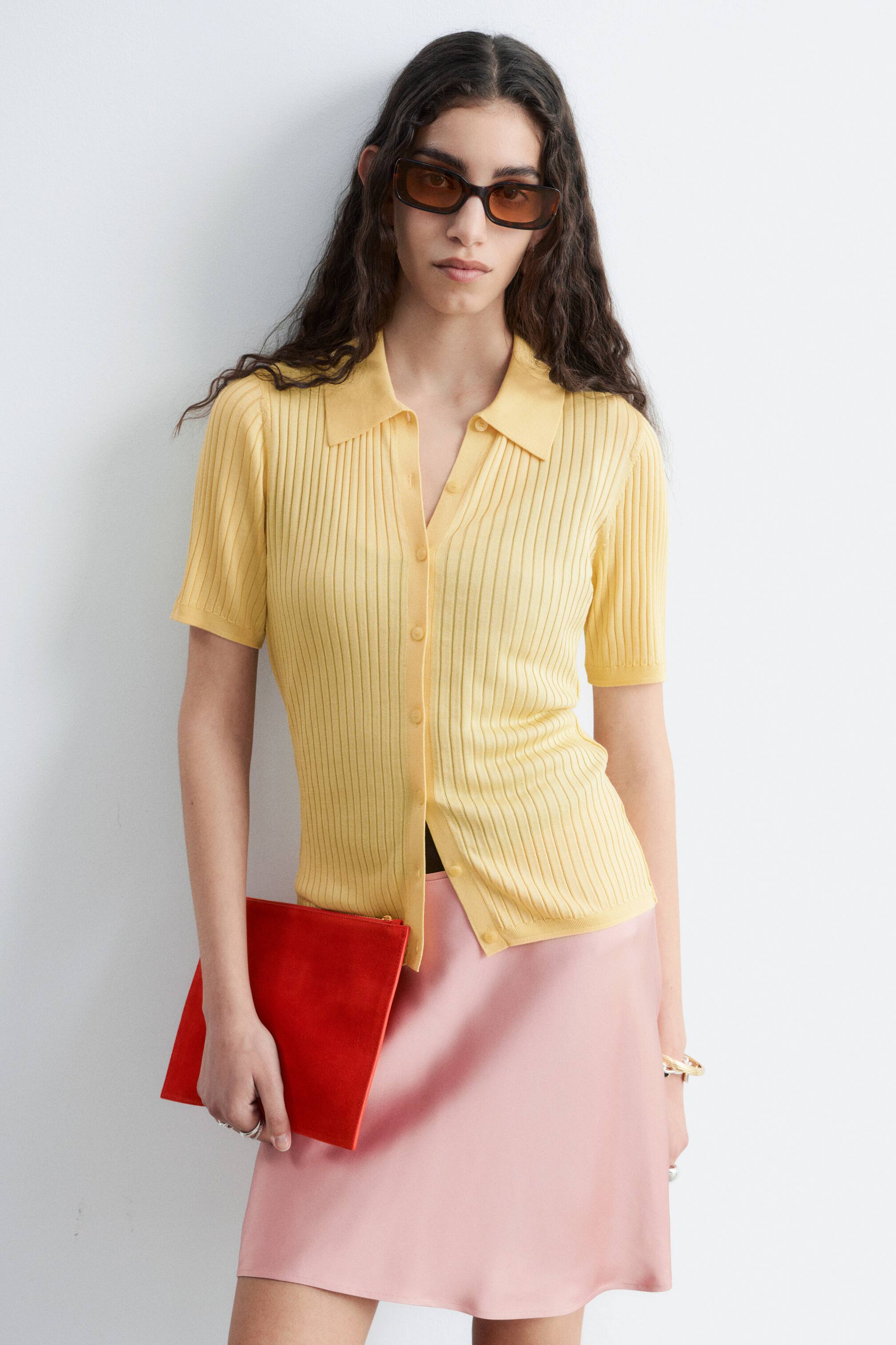 Zobacz większy obraz: Modelka ma na sobie żółty top z dzianiny prążkowanej z kołnierzykiem polo, krótkimi rękawami i zapięciem na guziki, który został zestawiony z różową spódnicą z kilkoma odpiętymi guzikami.