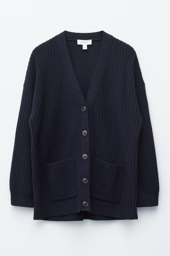 A dark blue ribbed cardigan is showcasing its FRONT, characterised by an oversized silhouette and a ribbed texture. A V-neckline forms at the top, with five tonal buttons running down the centre, and two patch pockets positioned on the lower front. - 1