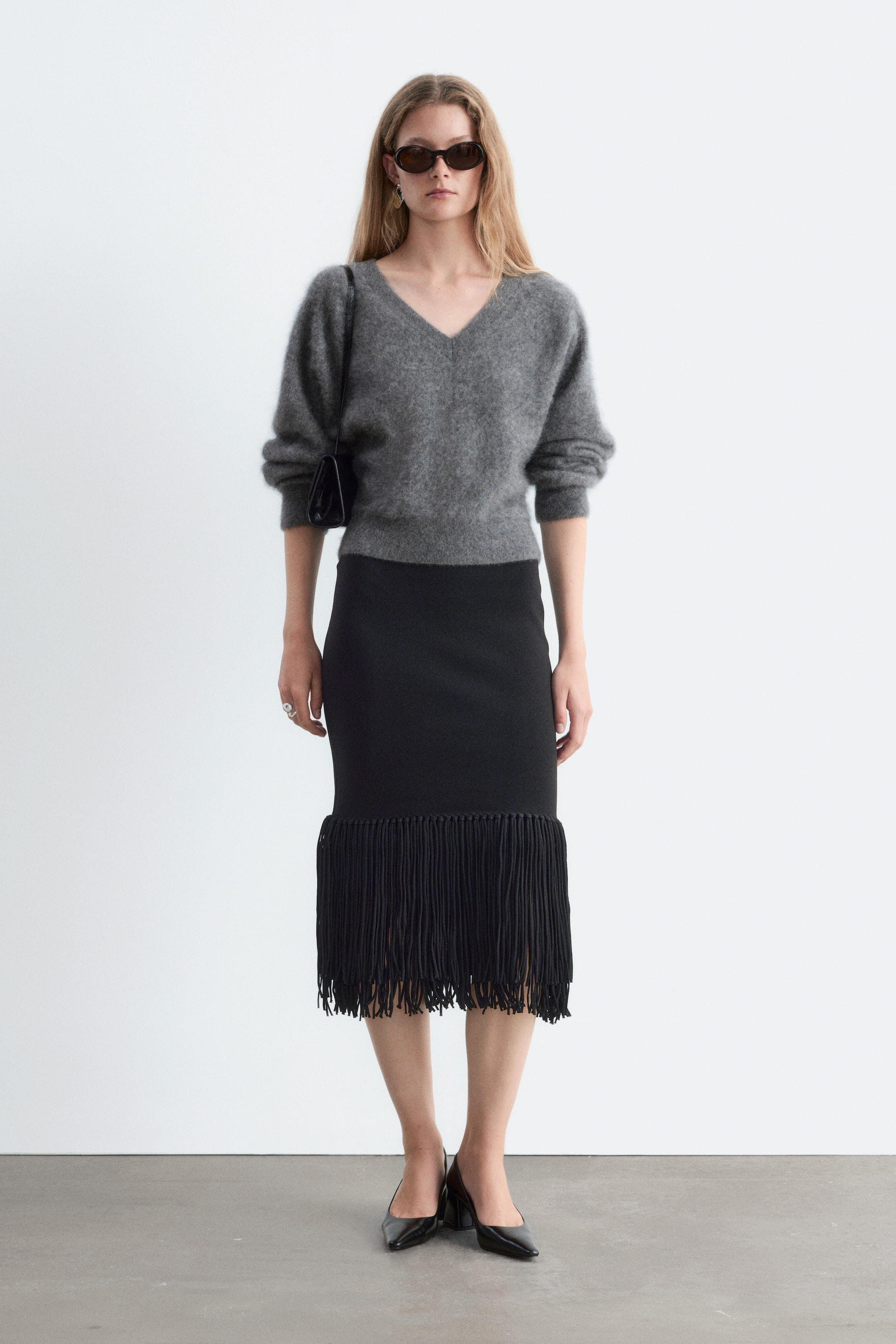 Größeres Bild ansehen: Ein schmaler schwarzer Midirock mit hoher Taille ist von vorn zu sehen und verfügt über eine gerade Silhouette, die in einem dicken, doppellagigen Fransensaum endet, der unterhalb der Knie fällt. Der Rock ist mit einem grauen Strickpullover mit langen Ärmeln kombiniert, der den Bund bedeckt.