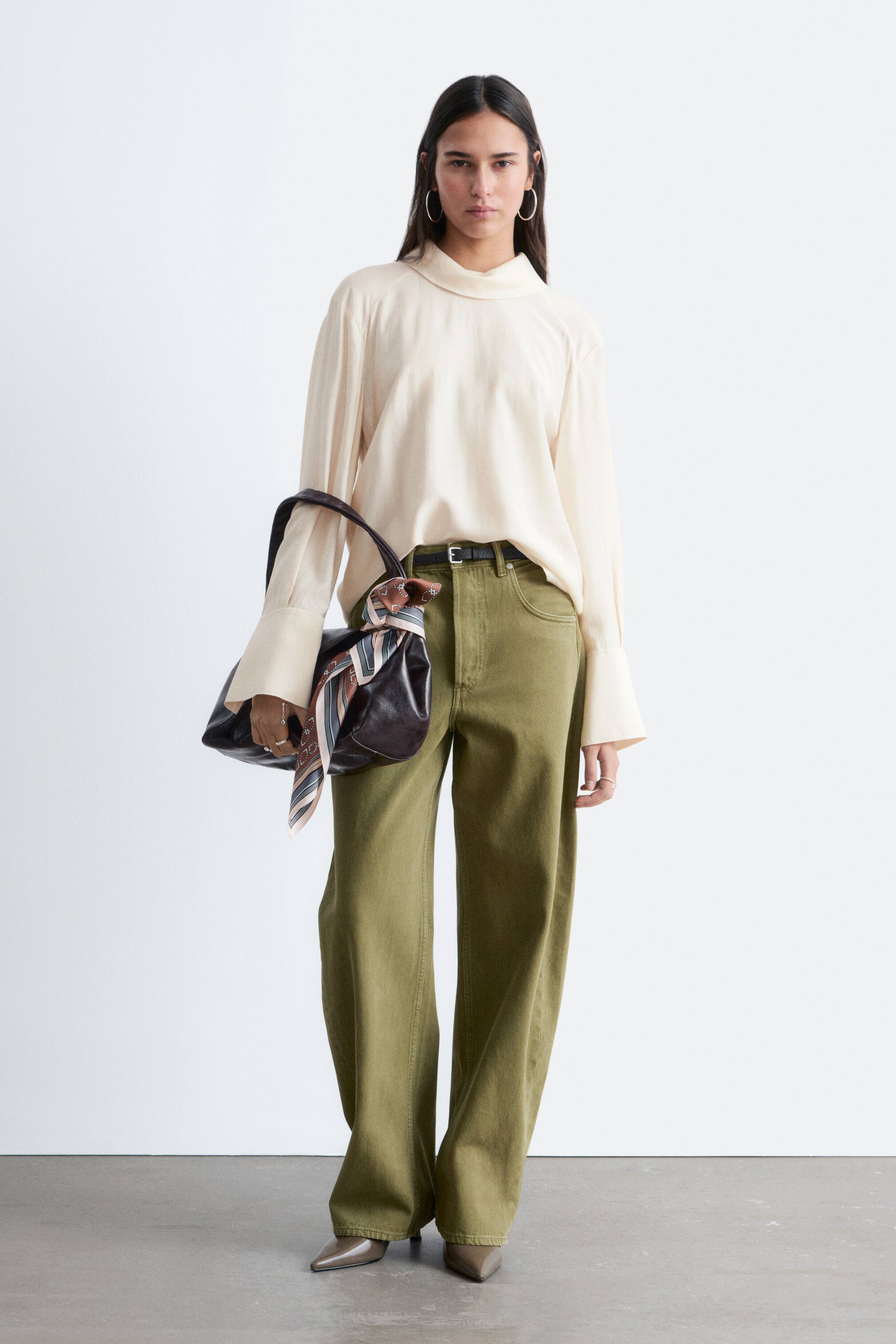 Agrandir l'image: Un ensemble orienté vers l'avant est présenté, mettant en vedette un chemisier à col montant blanc cassé avec des poignets larges, rentré dans un pantalon olive à taille haute et jambes larges, maintenu par une ceinture noire. Un sac à bandoulière marron foncé, orné d'une écharpe rayée, est porté. Des escarpins gris à bout pointu complètent le look.