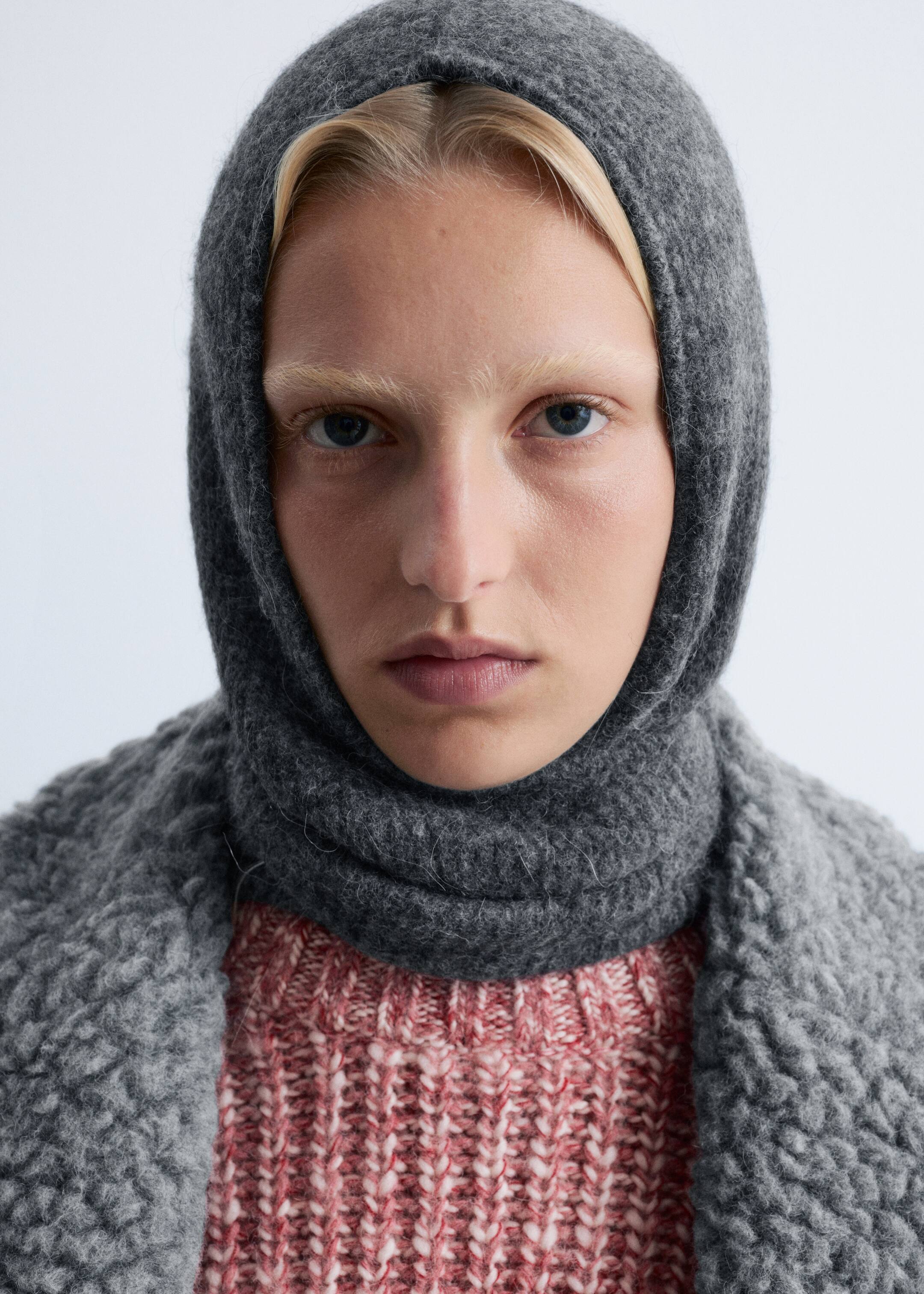 Größeres Bild anzeigen: Ein dunkelgraues Kapuzenteil ist von vorne zu sehen, bedeckt den Kopf und Hals und hat eine weiche, flauschige Strickstruktur. Es wird über einem pink-weißen groben Strickpullover getragen, mit einem hellgrauen, strukturierten Bouclé-Teil über die Schultern drapiert.