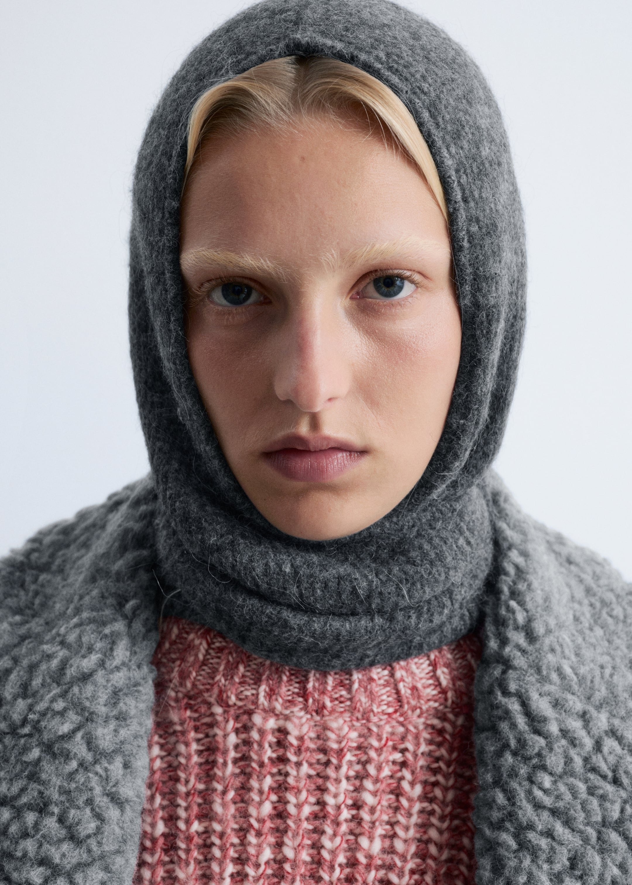 Größeres Bild ansehen: Ein dunkelgraues Kapuzenkleidungsstück ist in der Vorderansicht zu sehen, es bedeckt den Kopf und Hals und zeichnet sich durch eine weiche, flauschige Strickstruktur aus. Es wird über einem rosa-weiß gestreiften, grob gestrickten Pullover getragen, mit einem hellgrauen, strukturierten Bouclé-Kleidungsstück, das über die Schultern drapiert ist.
