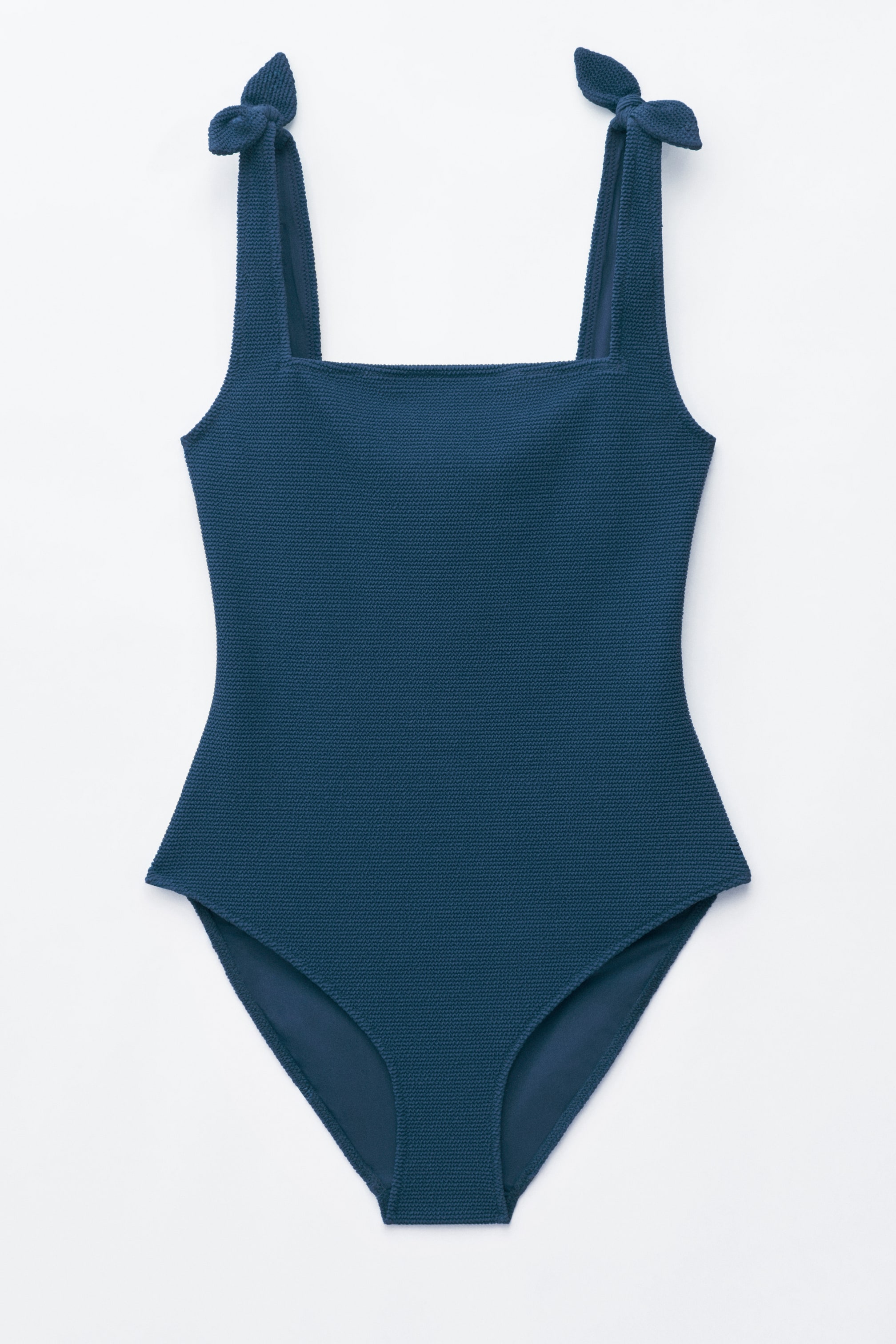 Visa större bild: En mörkblå texturerad baddräkt i ett stycke med fyrkantig halsringning och dekorativa rosetter vid axlarna, presenterad framifrån.