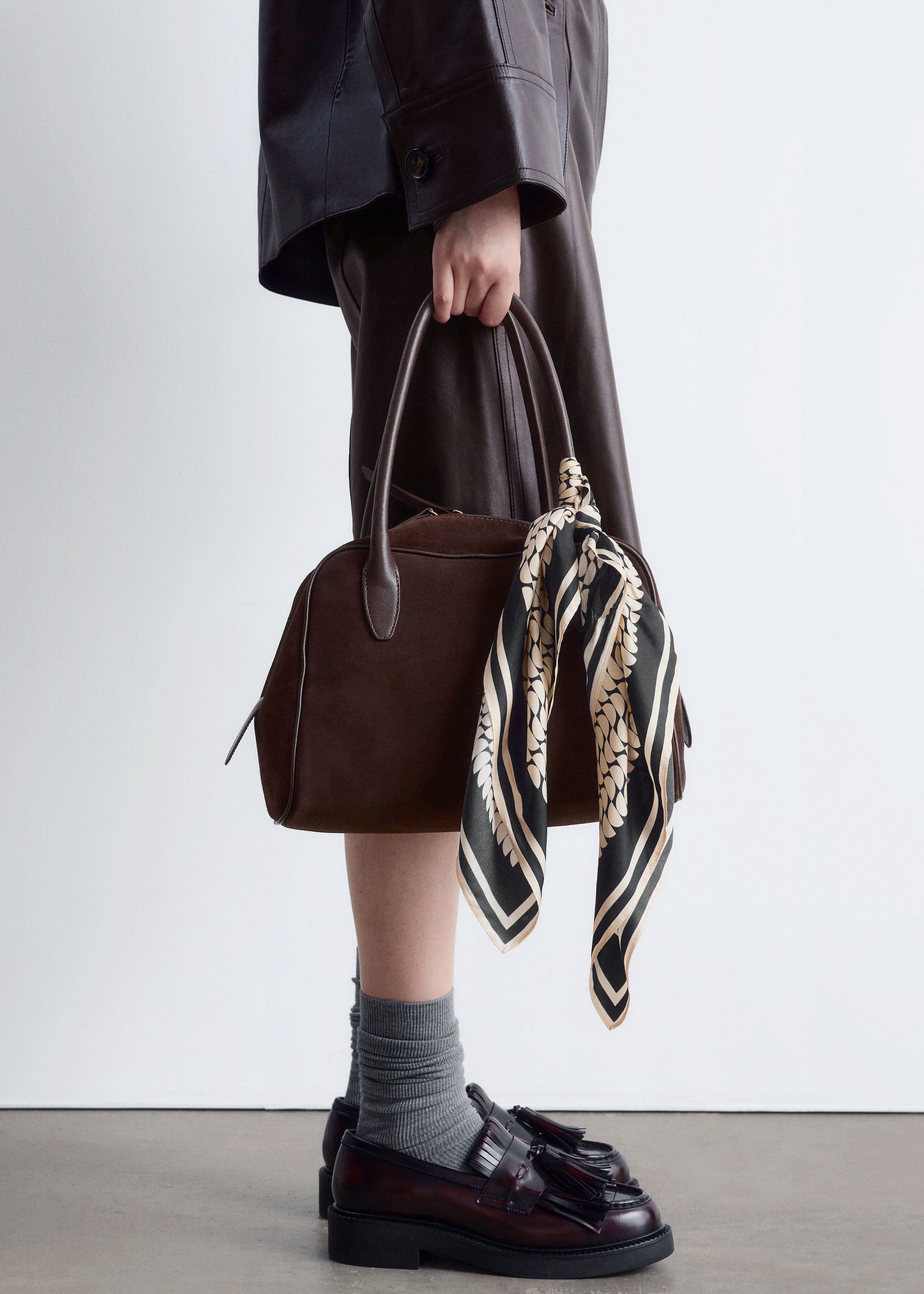 View larger image: A dark brown, suede-like handbag with smooth leather handles and piping is being held, with a black and cream patterned scarf tied to one handle. Below, dark red polished leather loafers, featuring tassel and kiltie details and chunky soles, are being worn with grey ribbed socks.