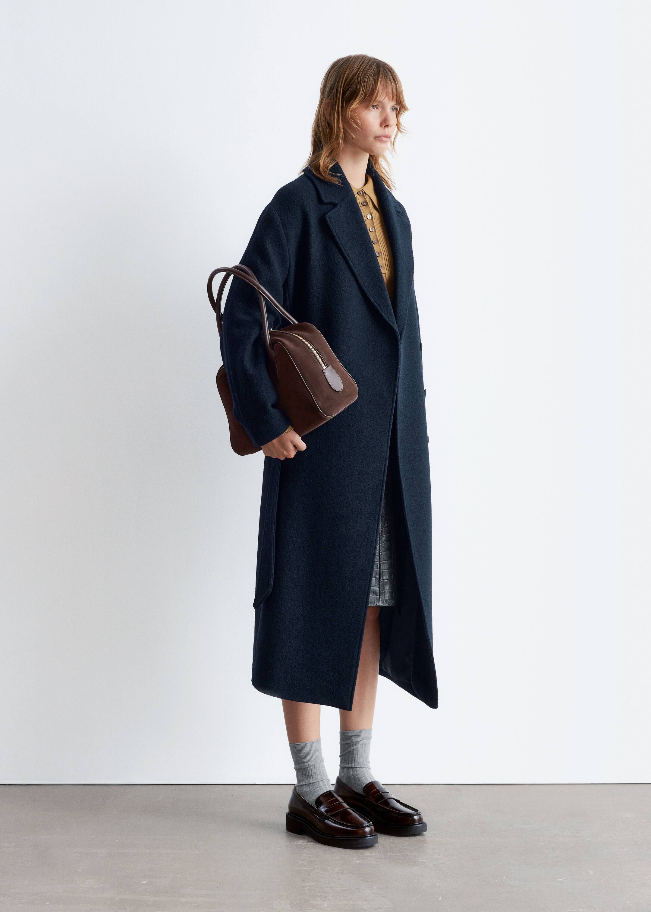 Agrandir l'image: Un manteau long bleu foncé est présenté de côté, mettant en valeur sa coupe décontractée et sa longueur complète, atteignant sous les genoux. Il présente des revers crantés, une poche plaquée visible et une ceinture à nouer pendante sur le côté gauche du porteur. Le manteau est porté ouvert, révélant une chemise à col jaune et une jupe grise à motifs en dessous.