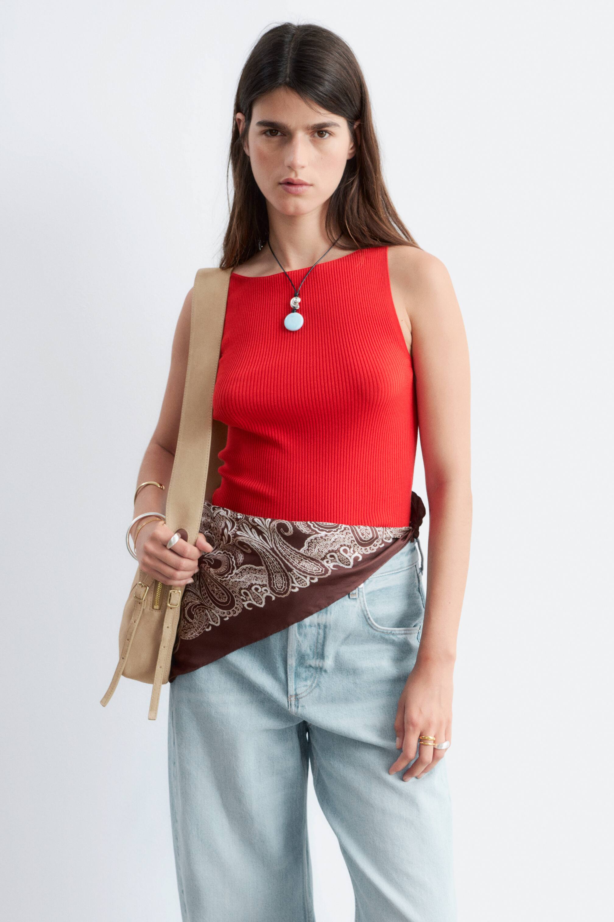 הצגת תמונה גדולה יותר: דגם לובשת גופיית סריג בצבע אדום עם מחשוף סירה. החולצה הצמודה מעוצבת עם שרשרת תליון בצבע כחול בהיר ומוכנסת לתוך ג'ינס בצבע שטיפה בהירה, עם צעיף פייזלי חום קשור סביב המותן.