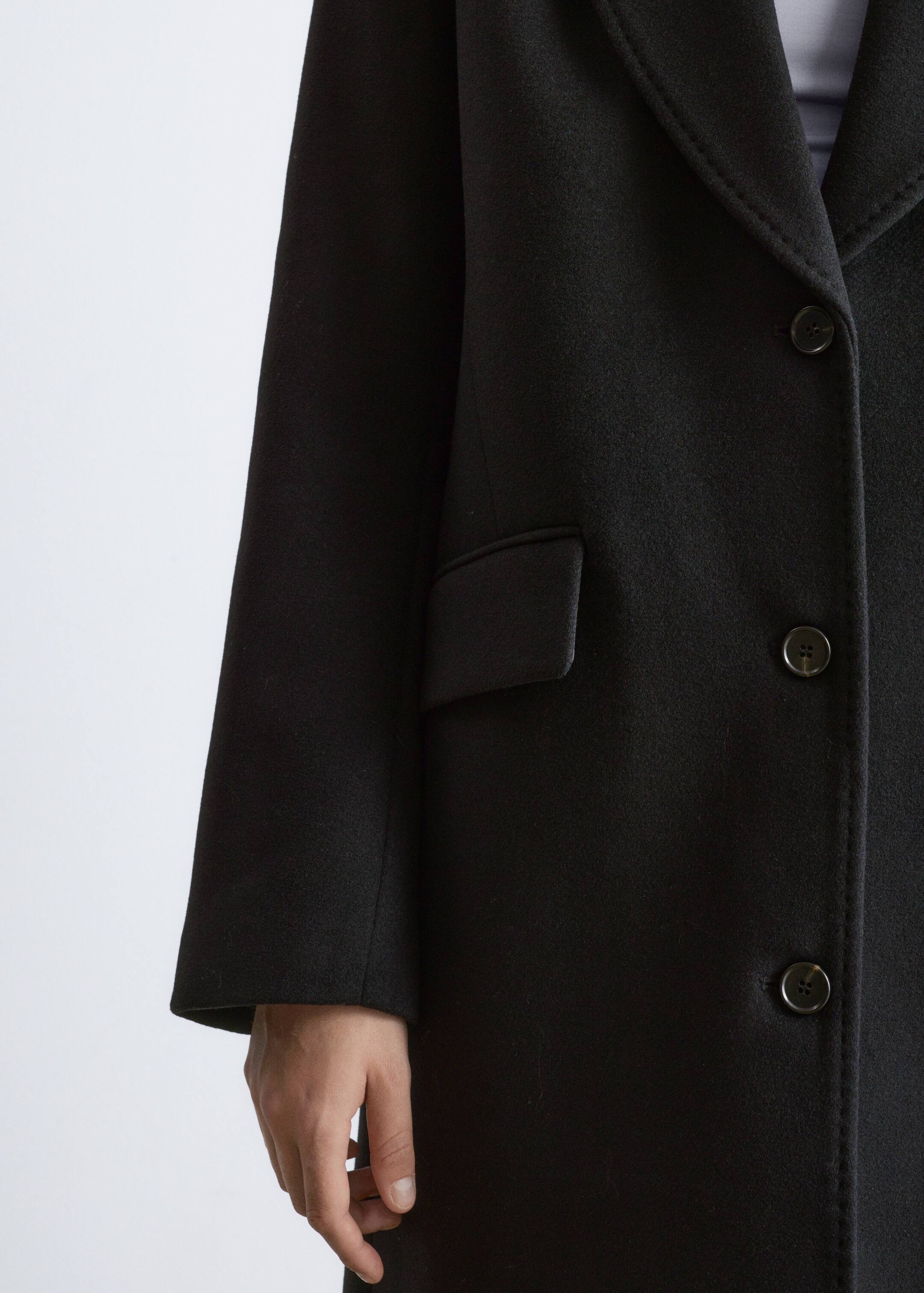 Agrandir l'image: Un manteau noir est présenté de face, avec des manches longues, un revers cranté, une poche à rabat sur le côté et une fermeture avant à trois boutons sombres. Une main sort de l'ouverture de la manche.