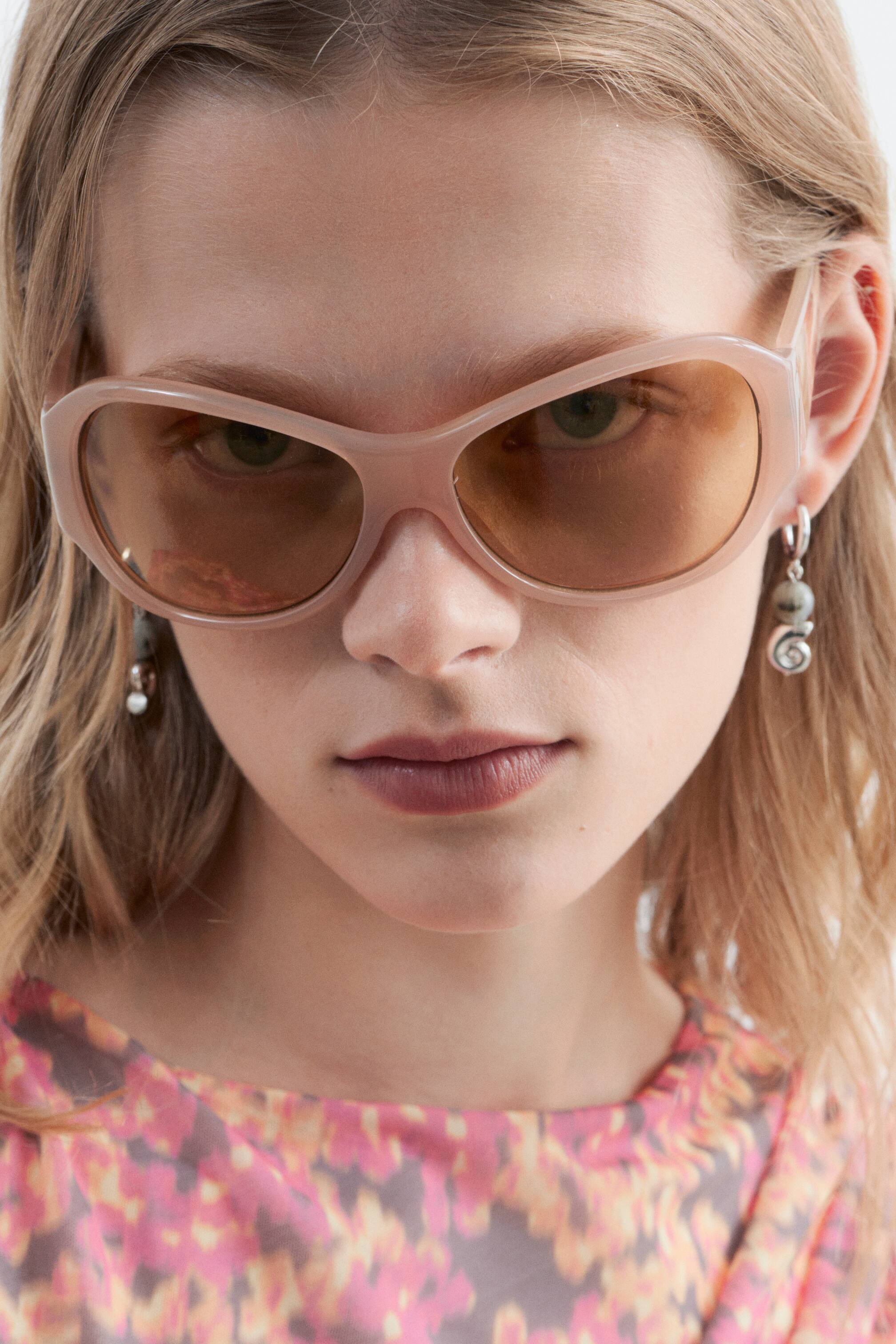 Größeres Bild ansehen: Hellrosa, übergroße Sonnenbrille mit gebogenen Acetatgestellen und braun getönten Gläsern werden von dem Model getragen. Die Augen des Models sind durch die Gläser sichtbar.