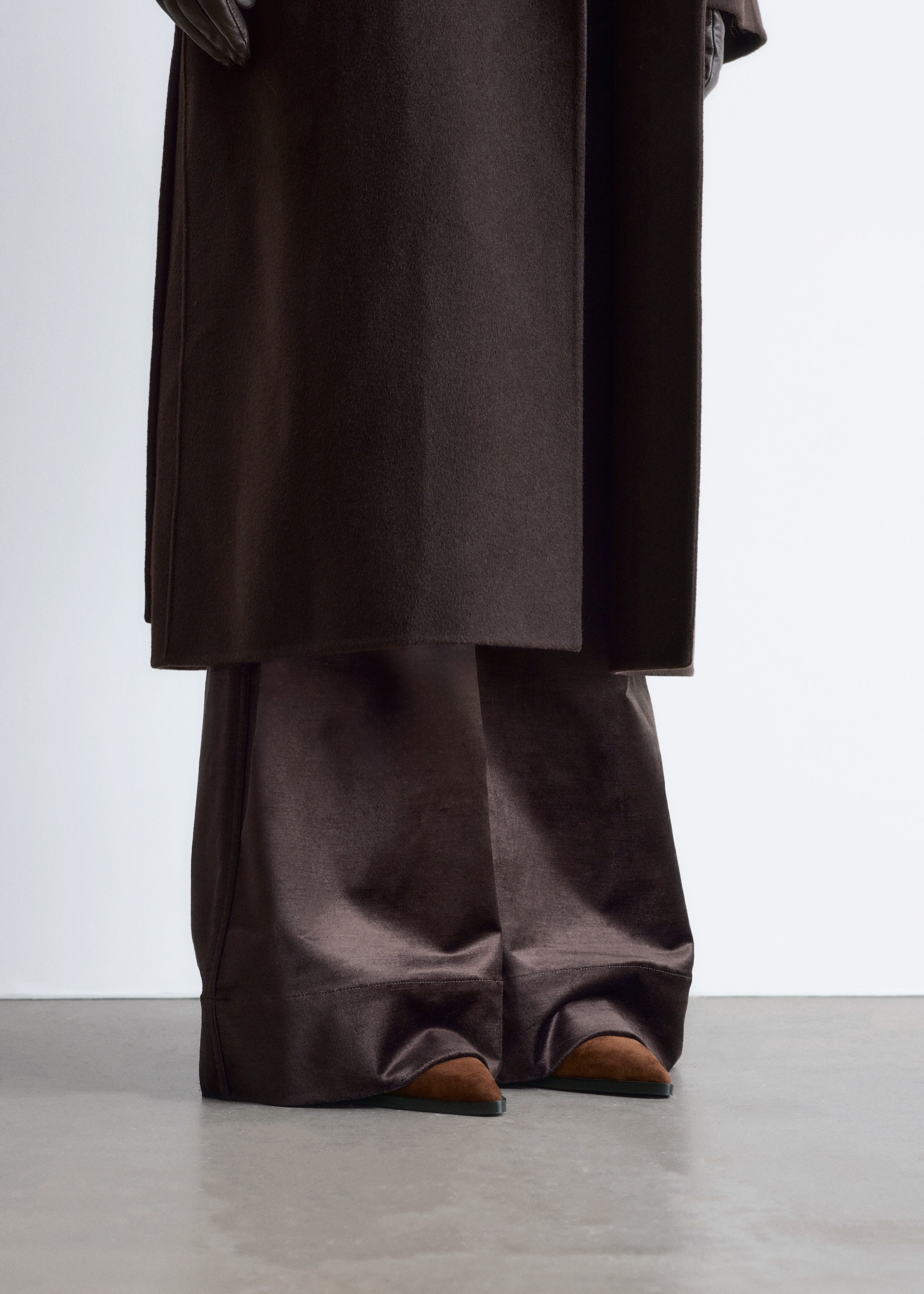 Visualizza immagine più grande: Un cappotto marrone scuro con texture opaca è stratificato su pantaloni a gamba larga e lucenti di colore marrone scuro. I pantaloni si arricciano sopra scarpe a punta, basse e di colore marrone rossiccio.